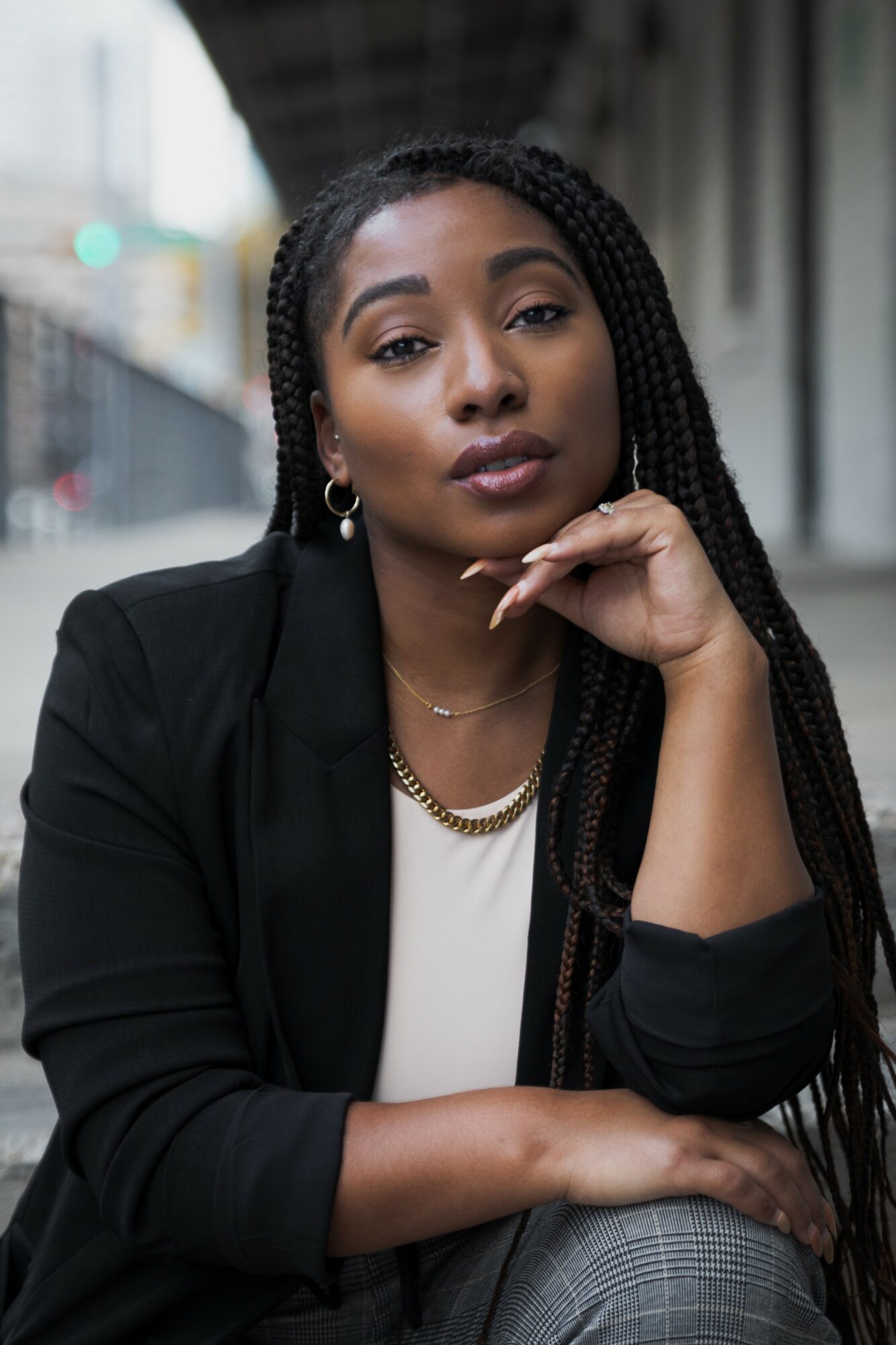 Woman with braided hair and earrings, resting her chin on her hand, outdoors in urban setting.