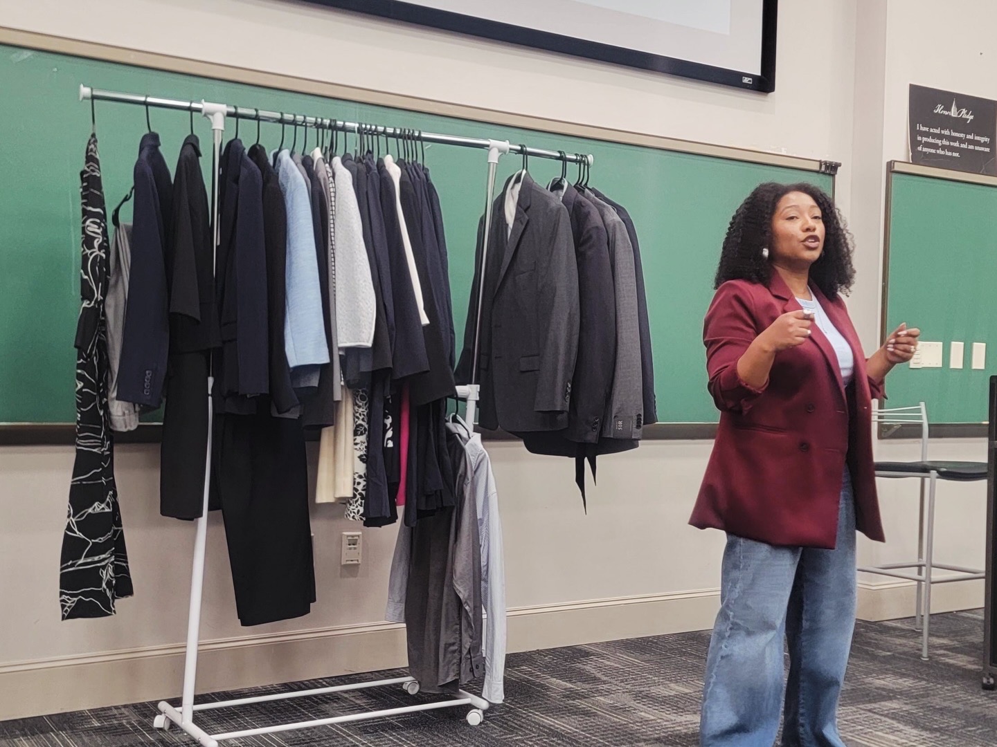 Woman standing in front of a clothing rack with various garments, in a classroom setting.