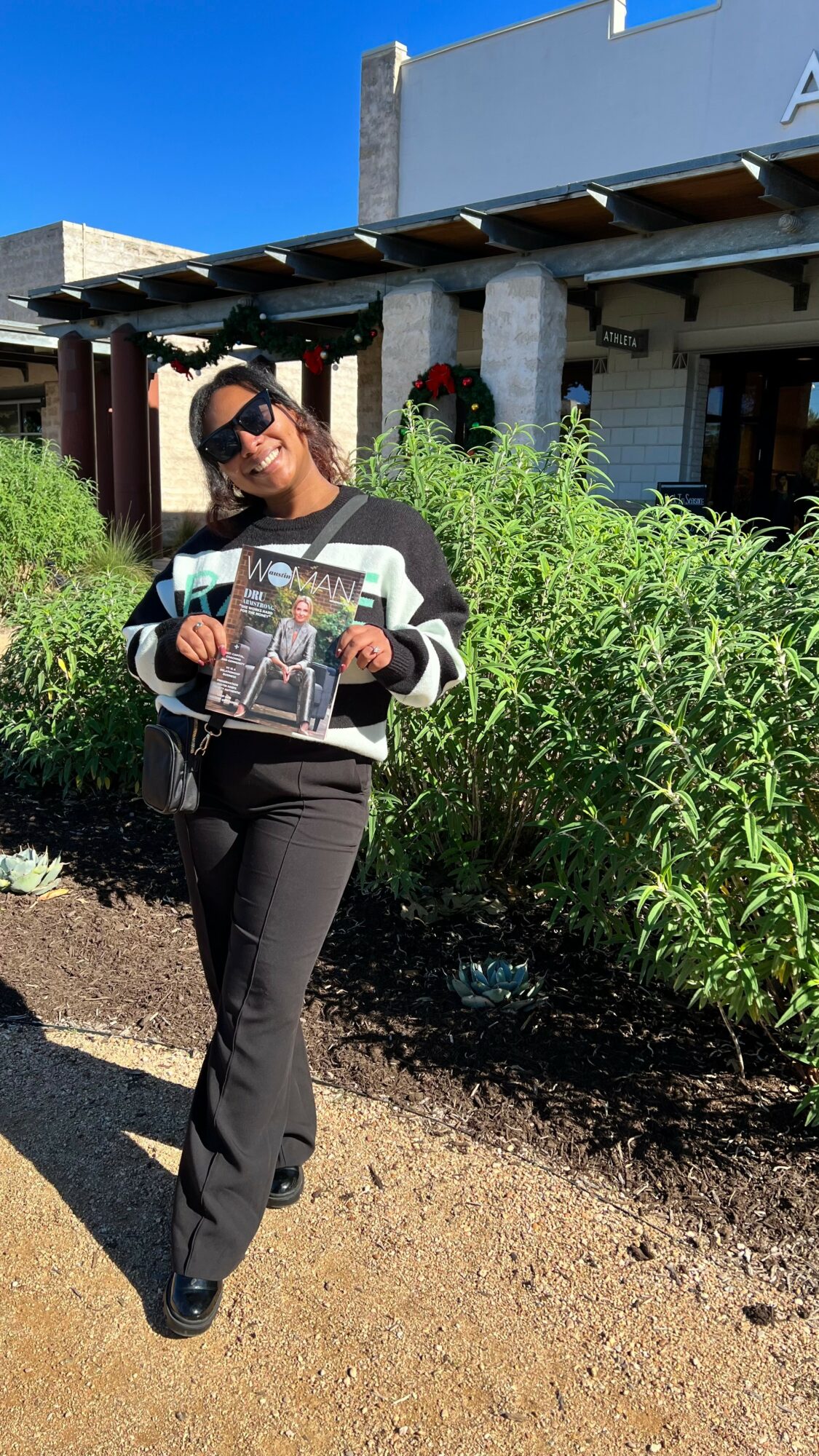 Woman standing outdoors holding a magazine, wearing sunglasses and a striped sweater, with greenery and a building in the background.