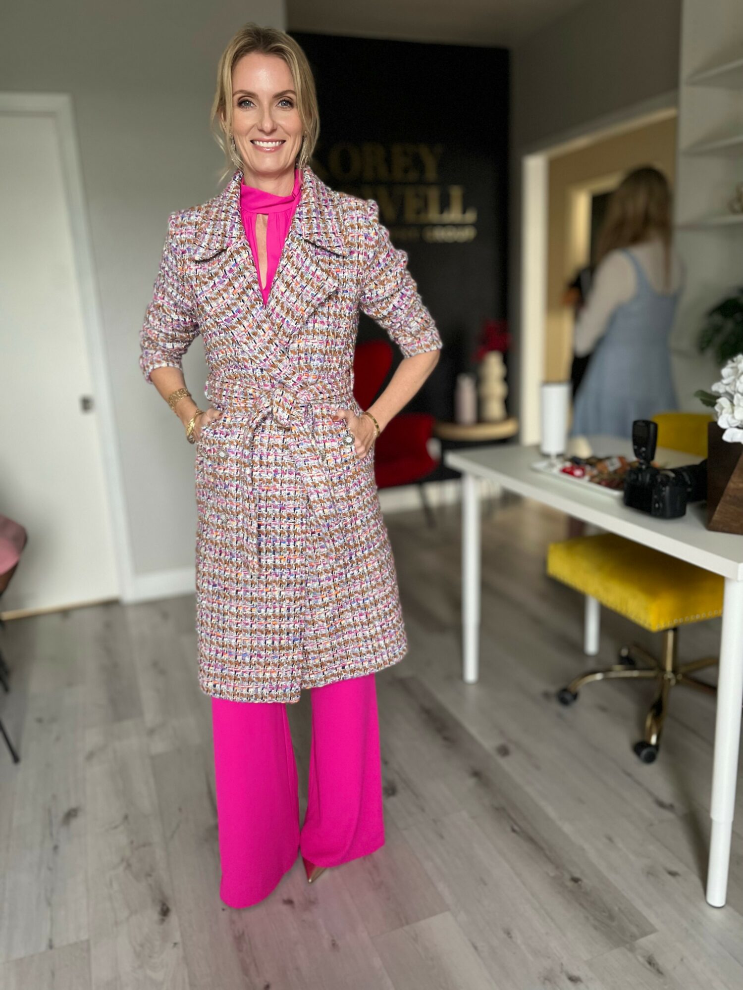 Woman in pink pants and patterned coat standing indoors, smiling, with a mirror and furniture in background.
