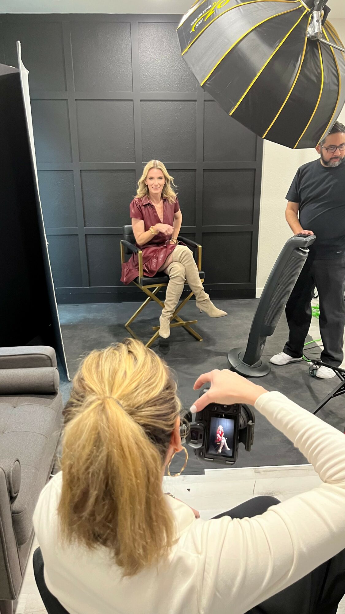 Woman sitting on a chair in front of a black wall, being photographed by a person with a camera, with a large studio light overhead.