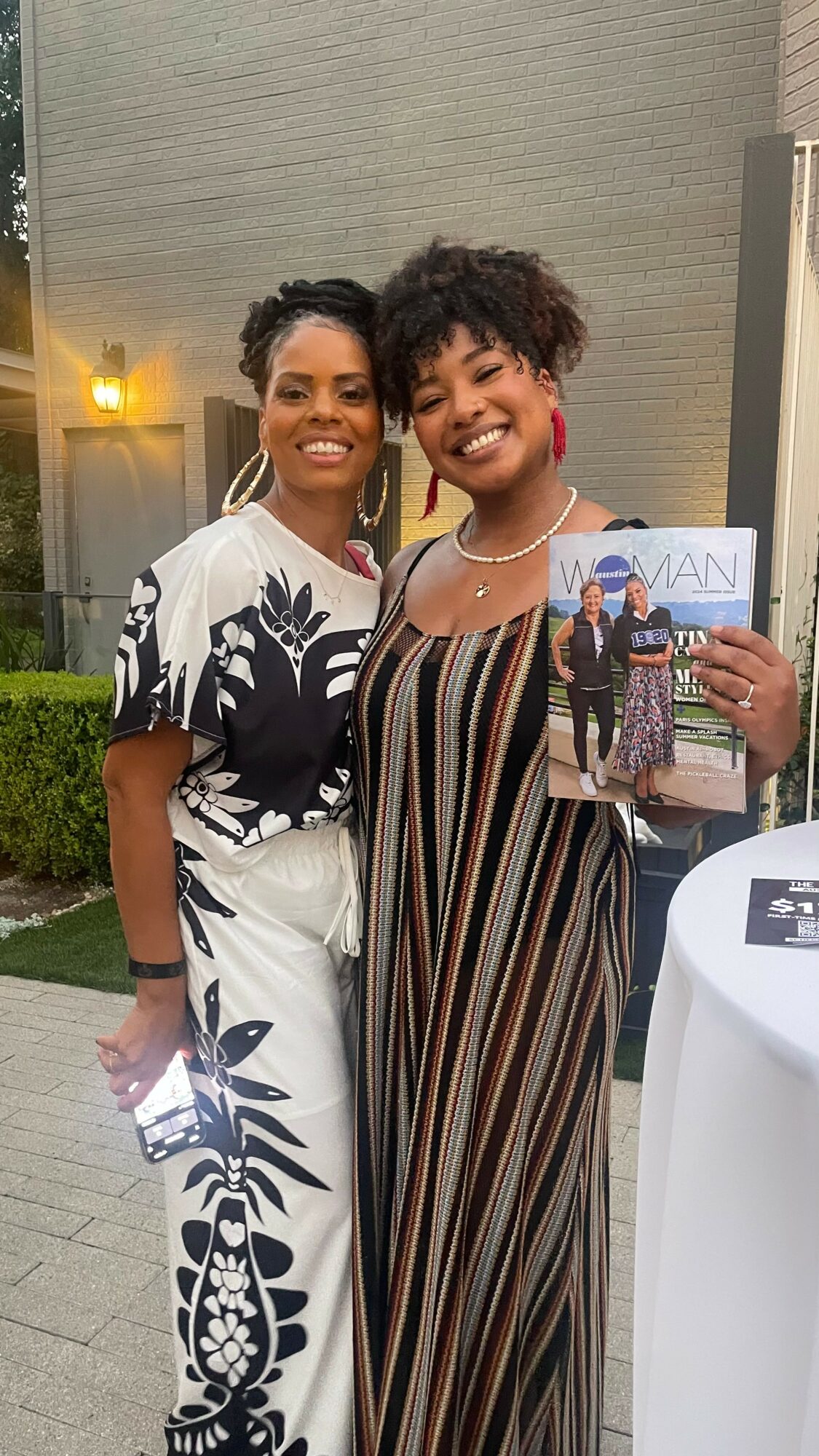 Two women smiling outdoors, one holding a magazine, with a building and greenery in the background.