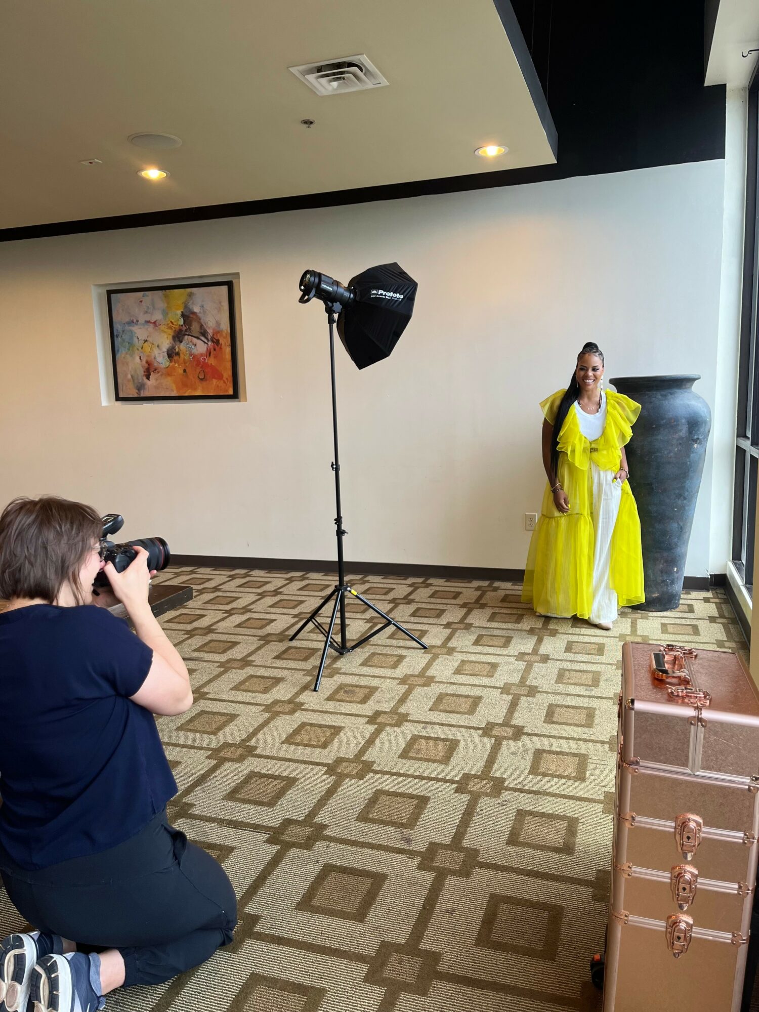 Person taking a photo of a woman in a yellow dress standing next to a large vase in a room with patterned carpet and wall art.