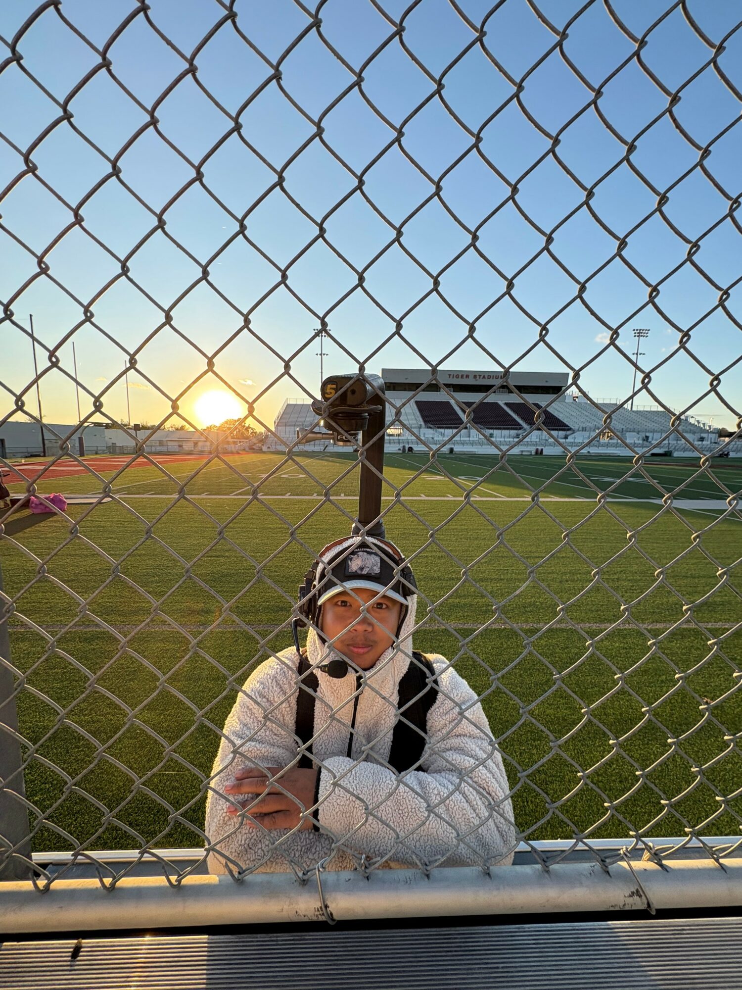 Child wearing a cap and jacket, leaning on a fence at a sports field during sunset.