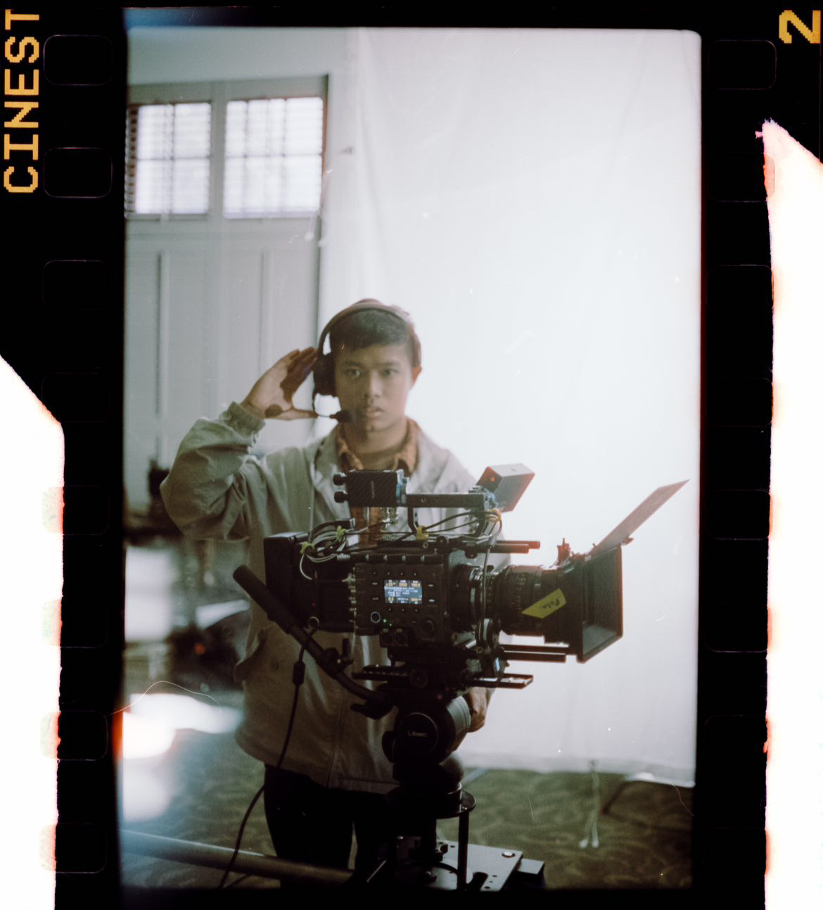 Person operating a camera on a tripod, holding a phone, in a room with windows and white walls.