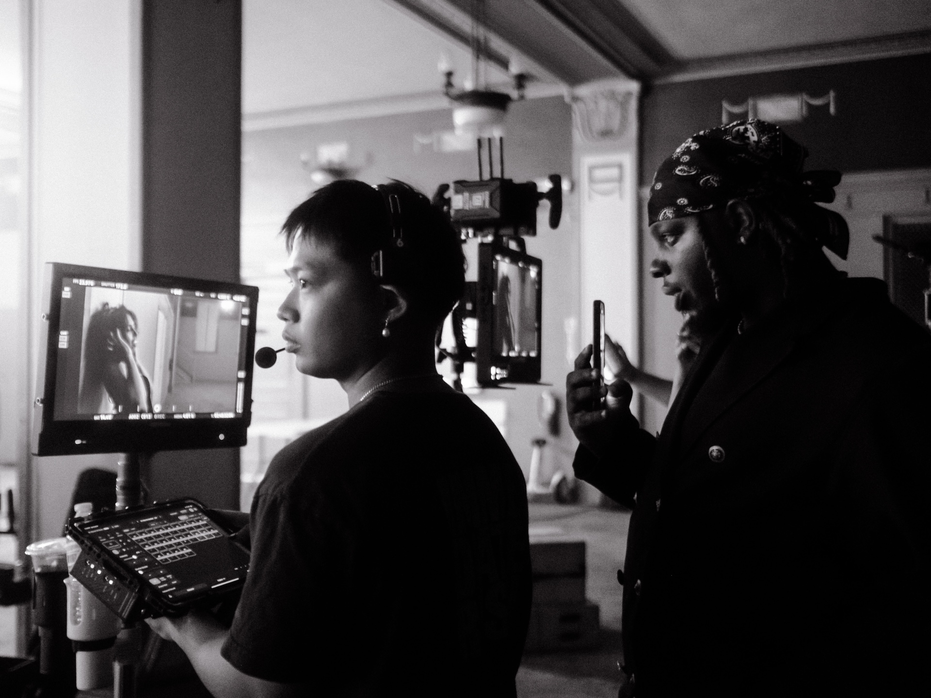 Two women working on a film set, one looking at a monitor and the other holding a device, in black and white.
