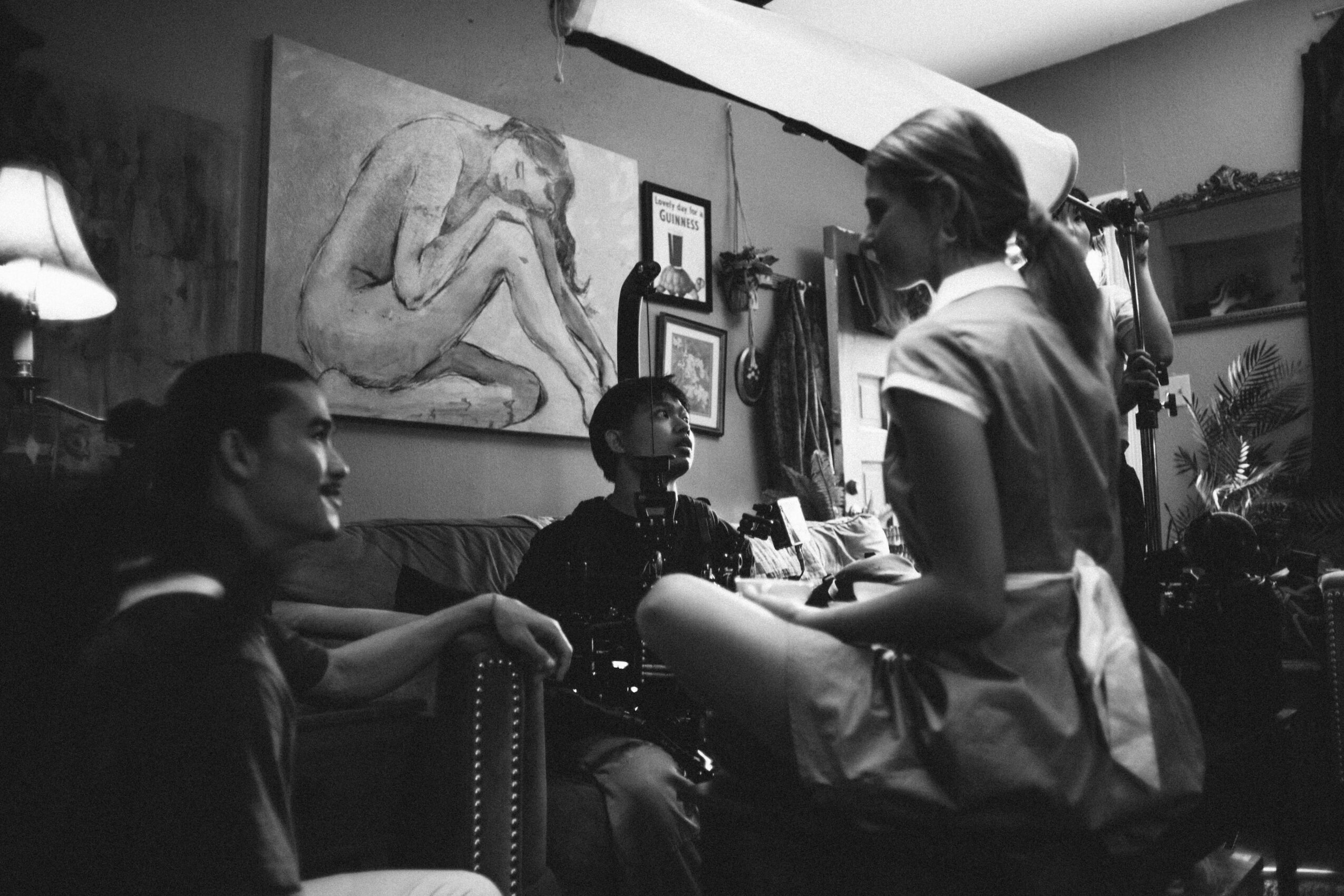 Three women in a room with artwork, one standing and two sitting, engaging in conversation, with shelves and decorations visible.