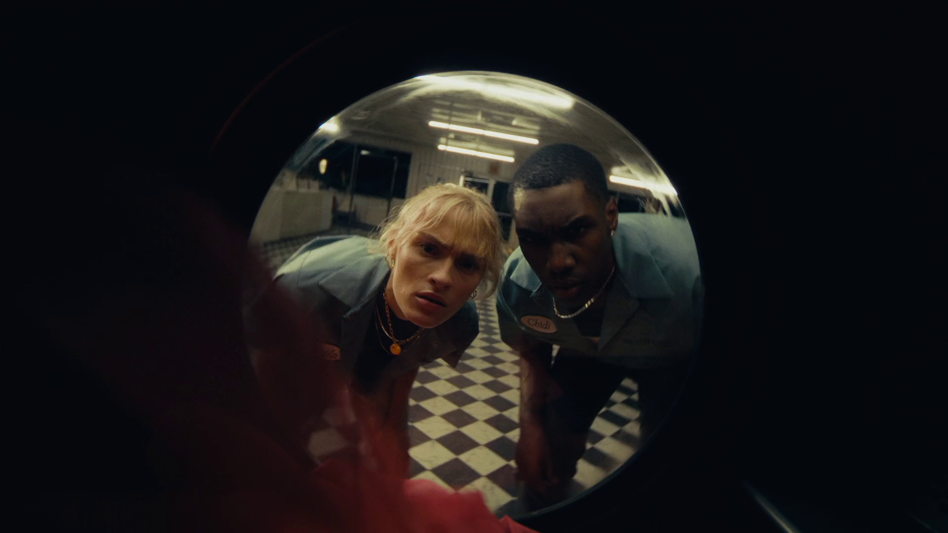 Two people looking into a circular mirror, with a checkered floor visible, in a room with fluorescent lighting.