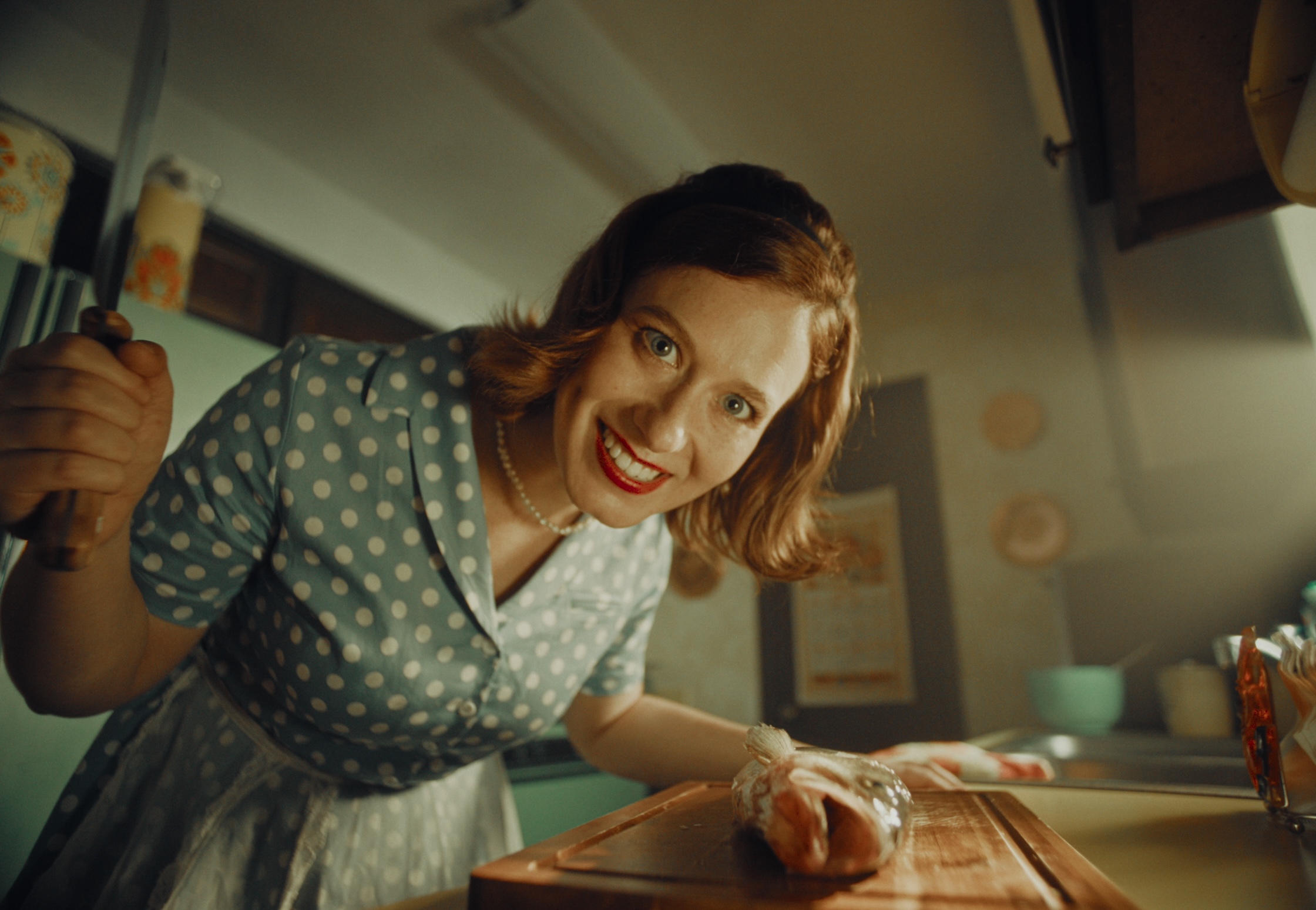 Woman with short hair in a polka dot dress leaning over a wooden cutting board, smiling at the camera.