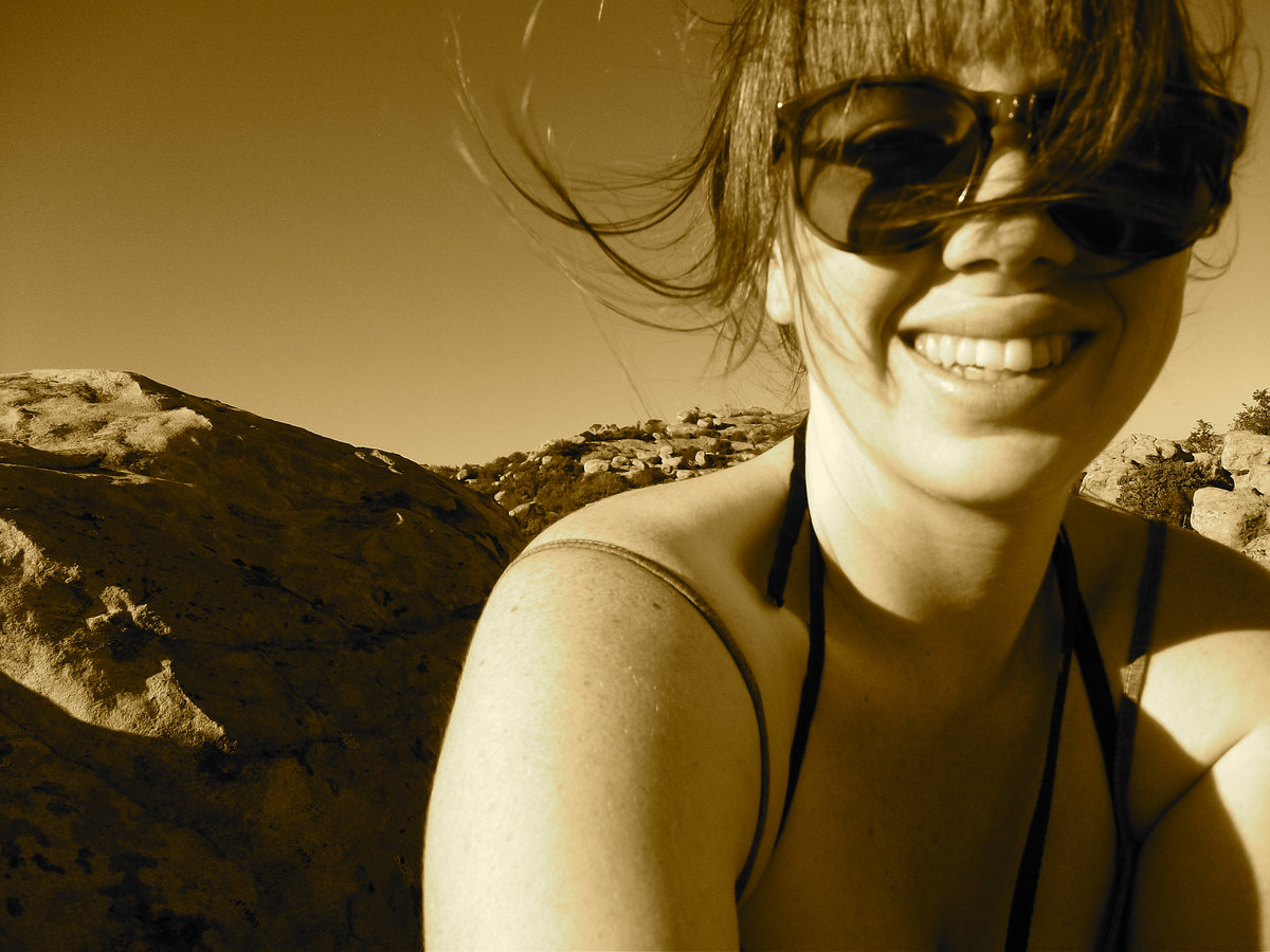 Smiling woman wearing sunglasses outdoors with rocky landscape in background.