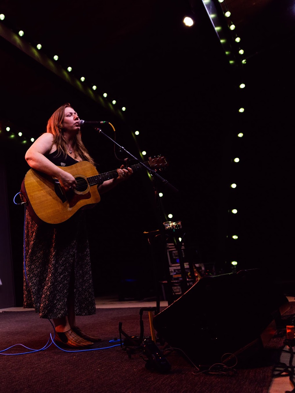A woman plays guitar and sings into a microphone on stage with string lights overhead.