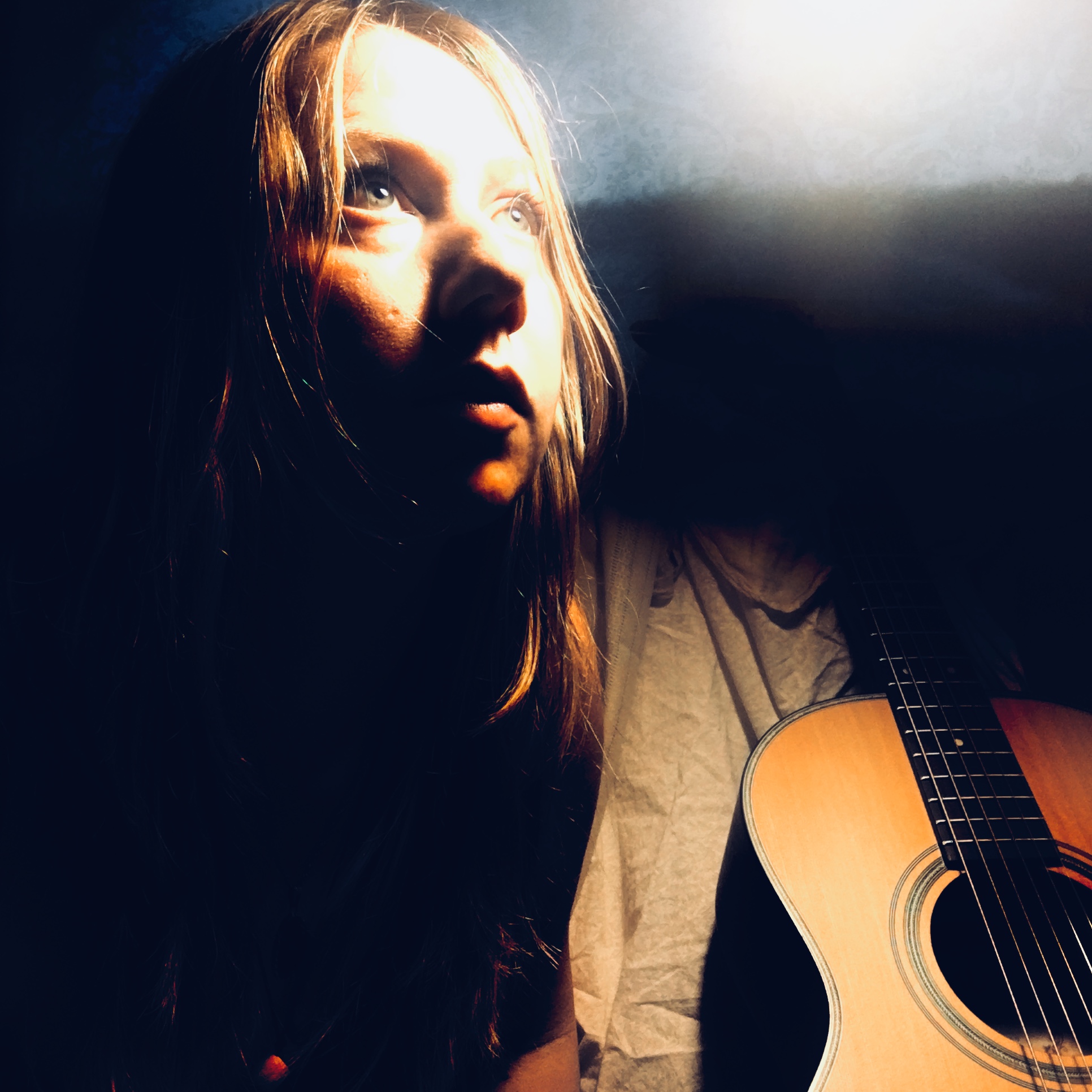 Young woman with long hair looking upward, illuminated by a bright light, with a guitar partially visible beside her.