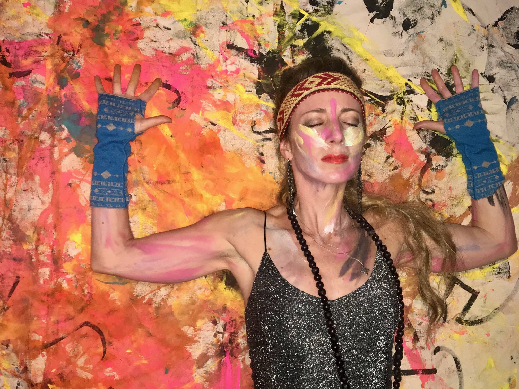 Woman with painted face and jewelry, arms raised, colorful abstract background, expressive pose.