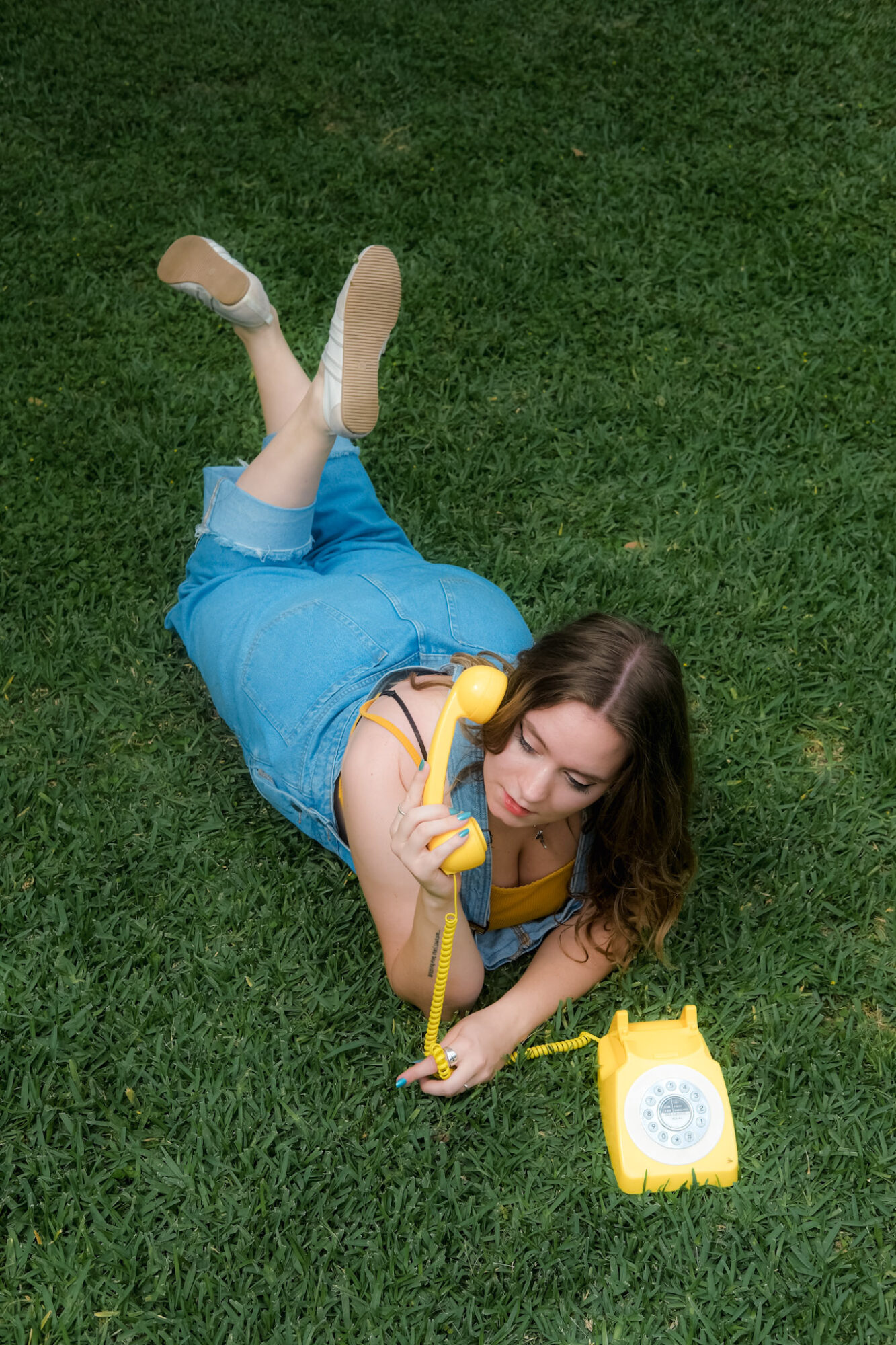 Young woman lying on grass, holding a yellow rotary phone, with a yellow phone base nearby, smiling.