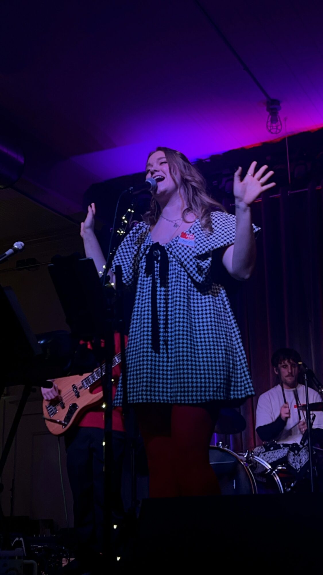 A woman singing on stage with a microphone, wearing a checkered dress and gesturing with her hands, with musicians in the background.