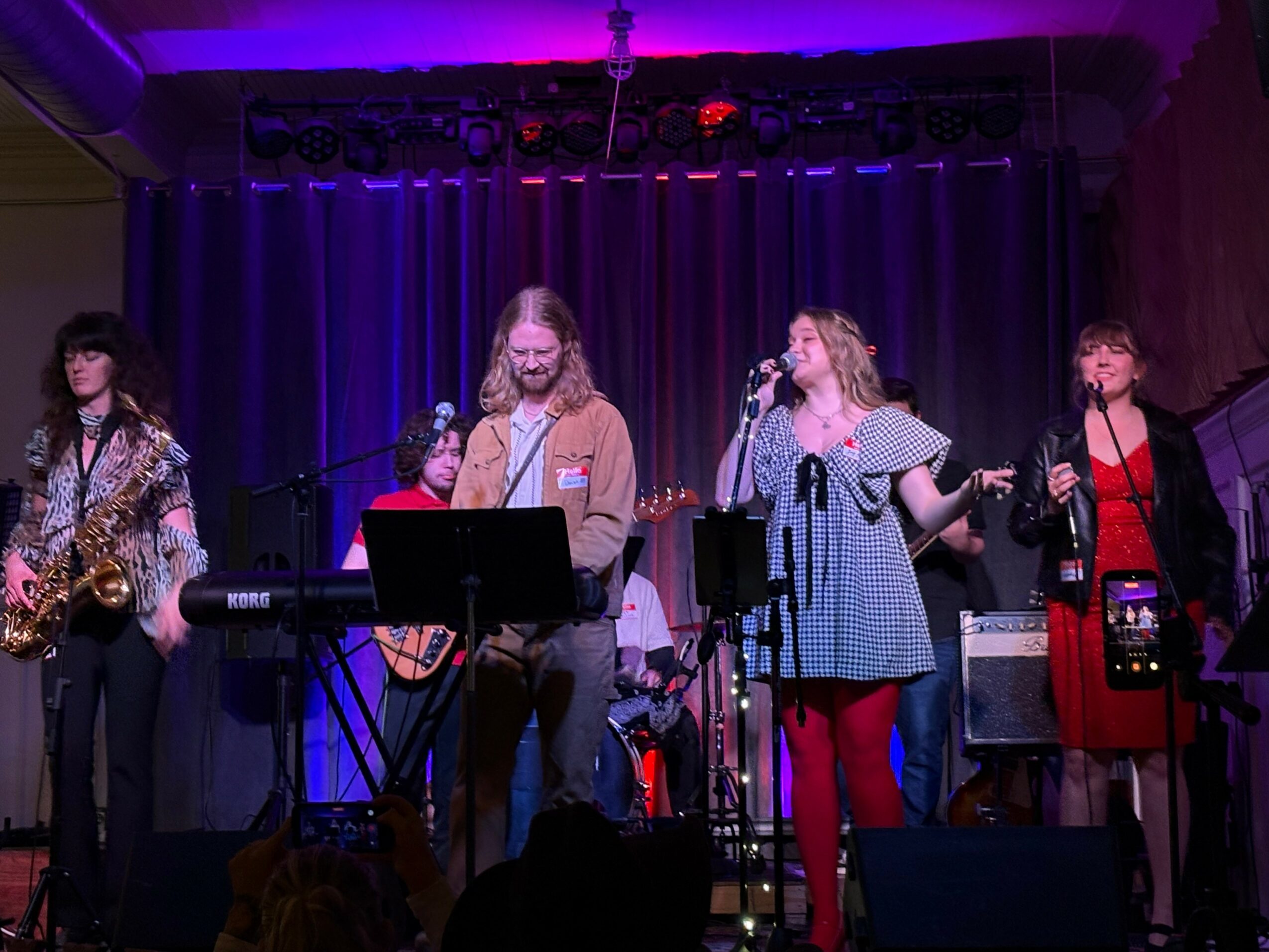 Group of five women performing on stage with musical instruments and microphones, purple lighting, curtain backdrop.