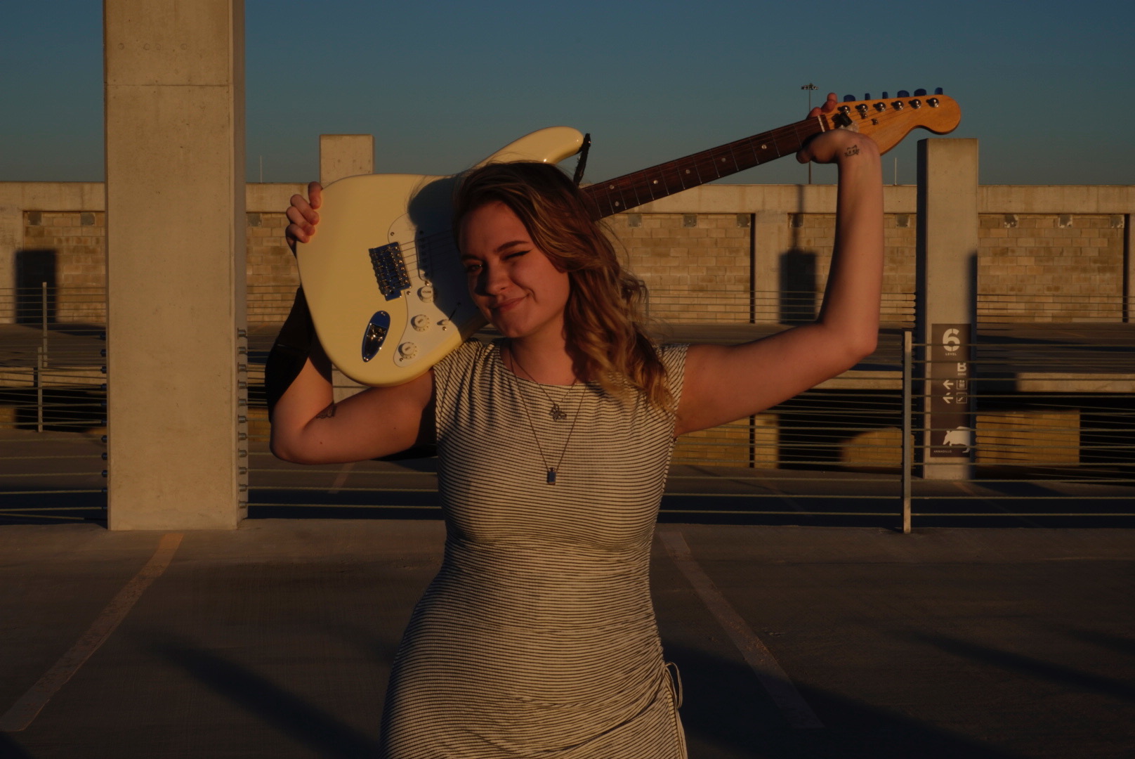 Young woman holding a guitar over her shoulder in an outdoor parking lot during sunset.