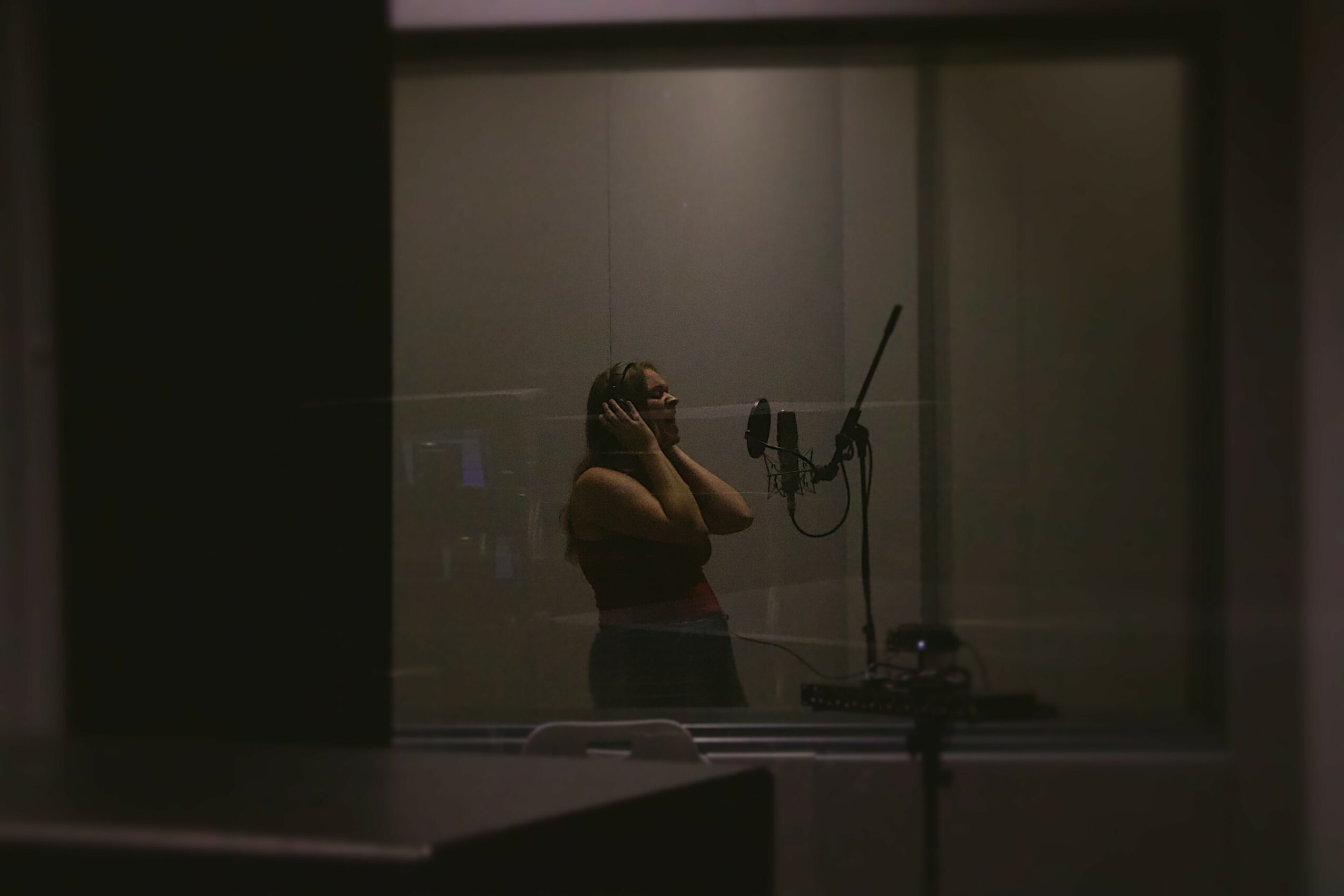 Person in a recording studio singing into a microphone, with headphones on, seen through a window.