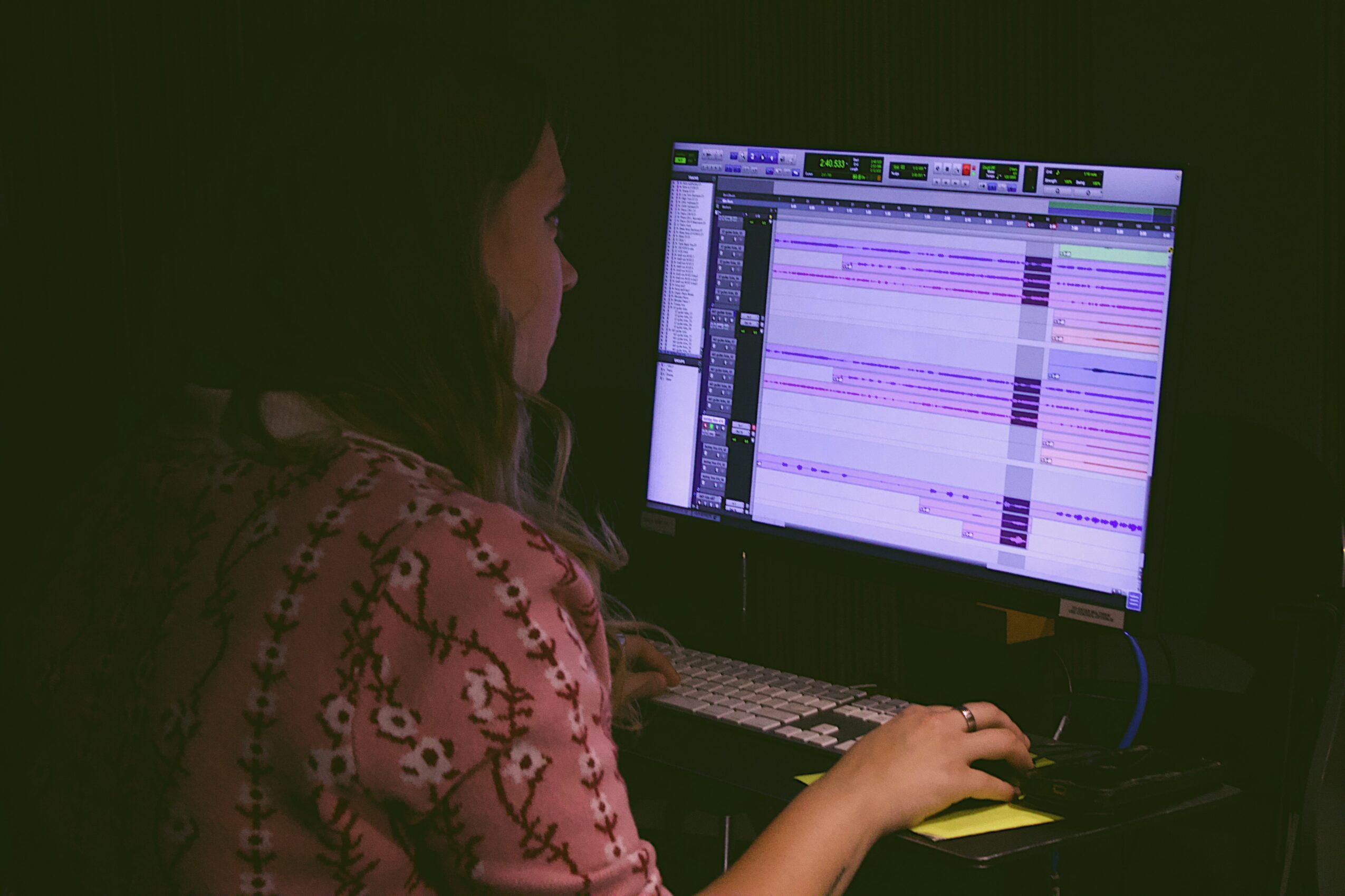 Person working on a computer with a digital audio workstation interface displayed on the screen, in a dark room.