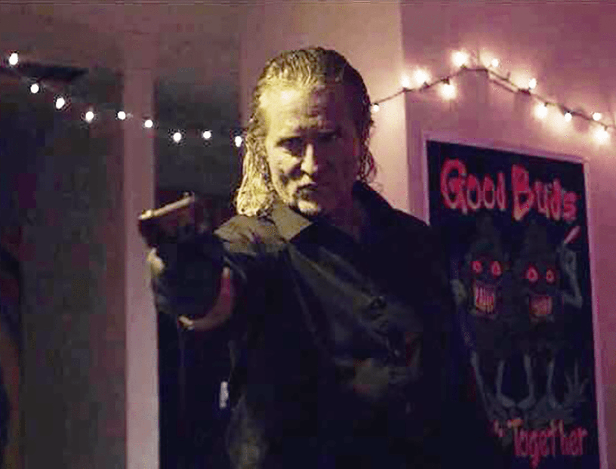 Person with long hair pointing a finger, dimly lit room with string lights and a colorful poster in the background.