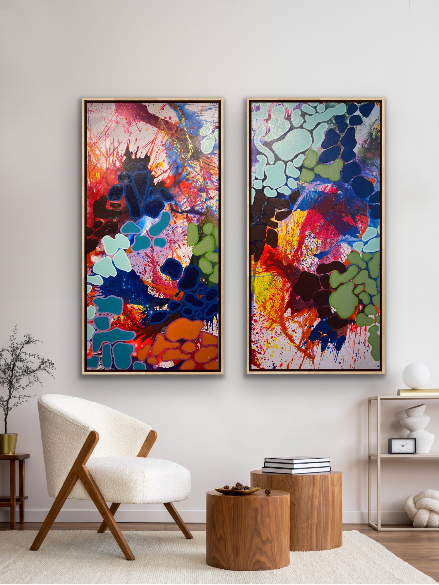 Two colorful abstract paintings hang on a white wall above a white armchair with wooden legs, two wooden stools, and a small shelf.