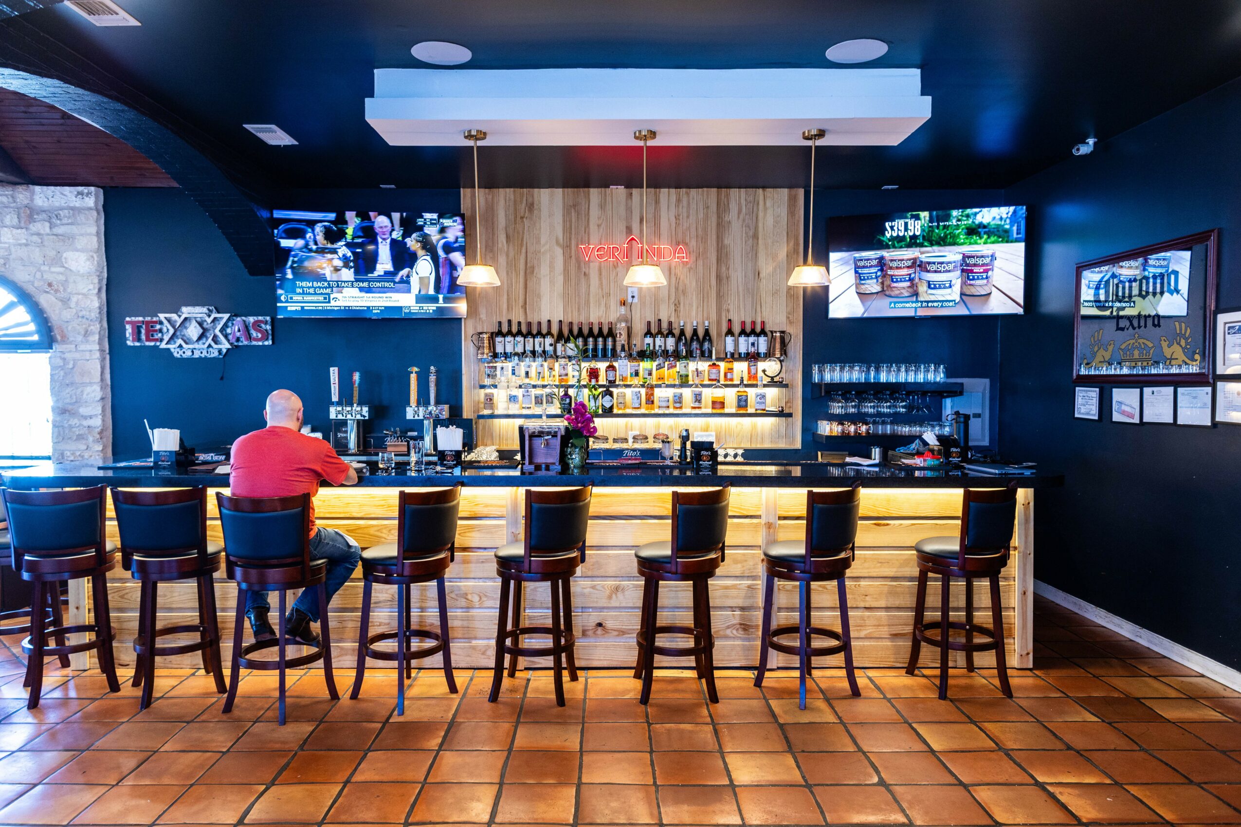 Bar with a person sitting on a stool, shelves with bottles, TVs on walls, and a tiled floor.