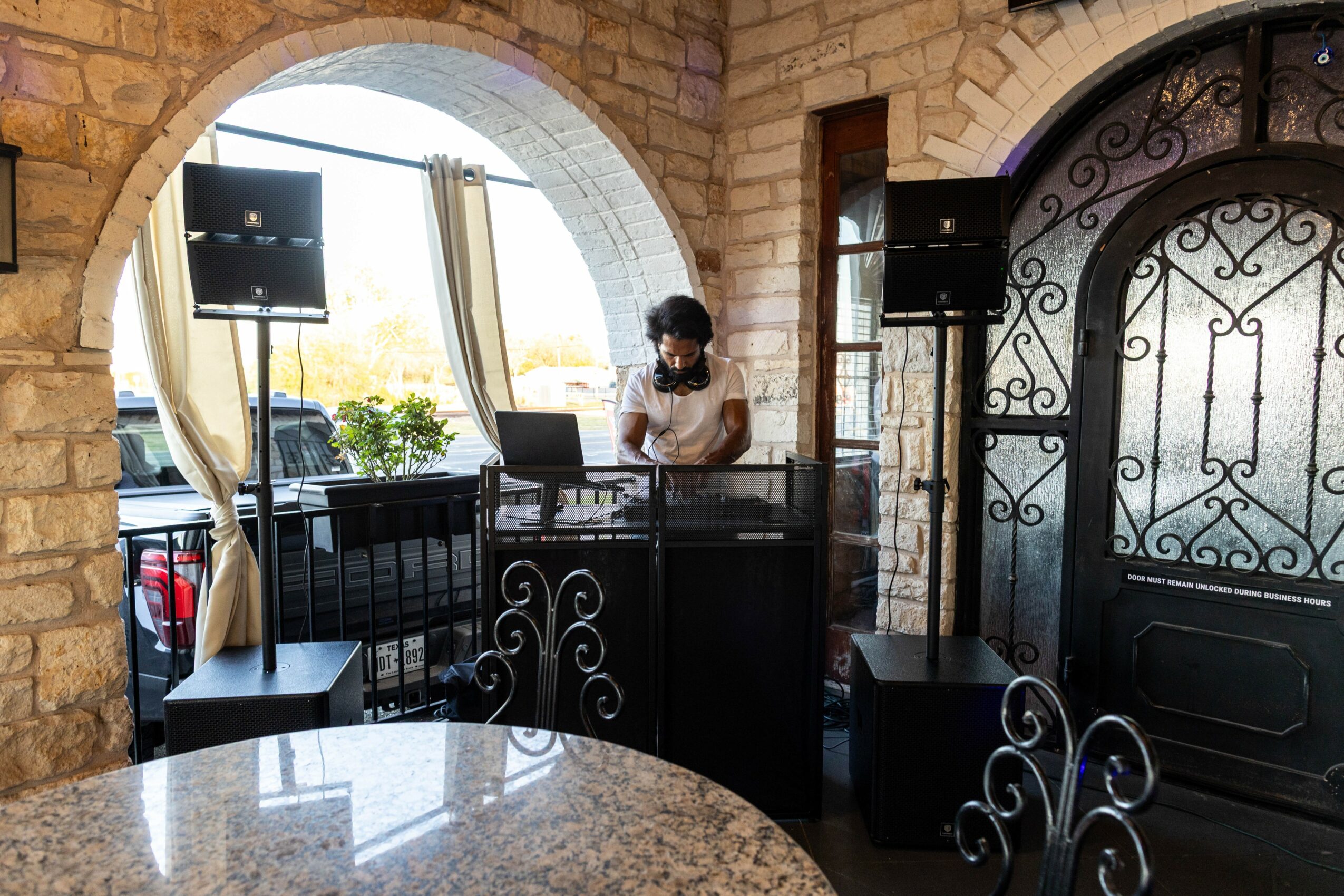 Person wearing headphones at a DJ booth inside a brick-walled room with large arched windows and decorative ironwork.
