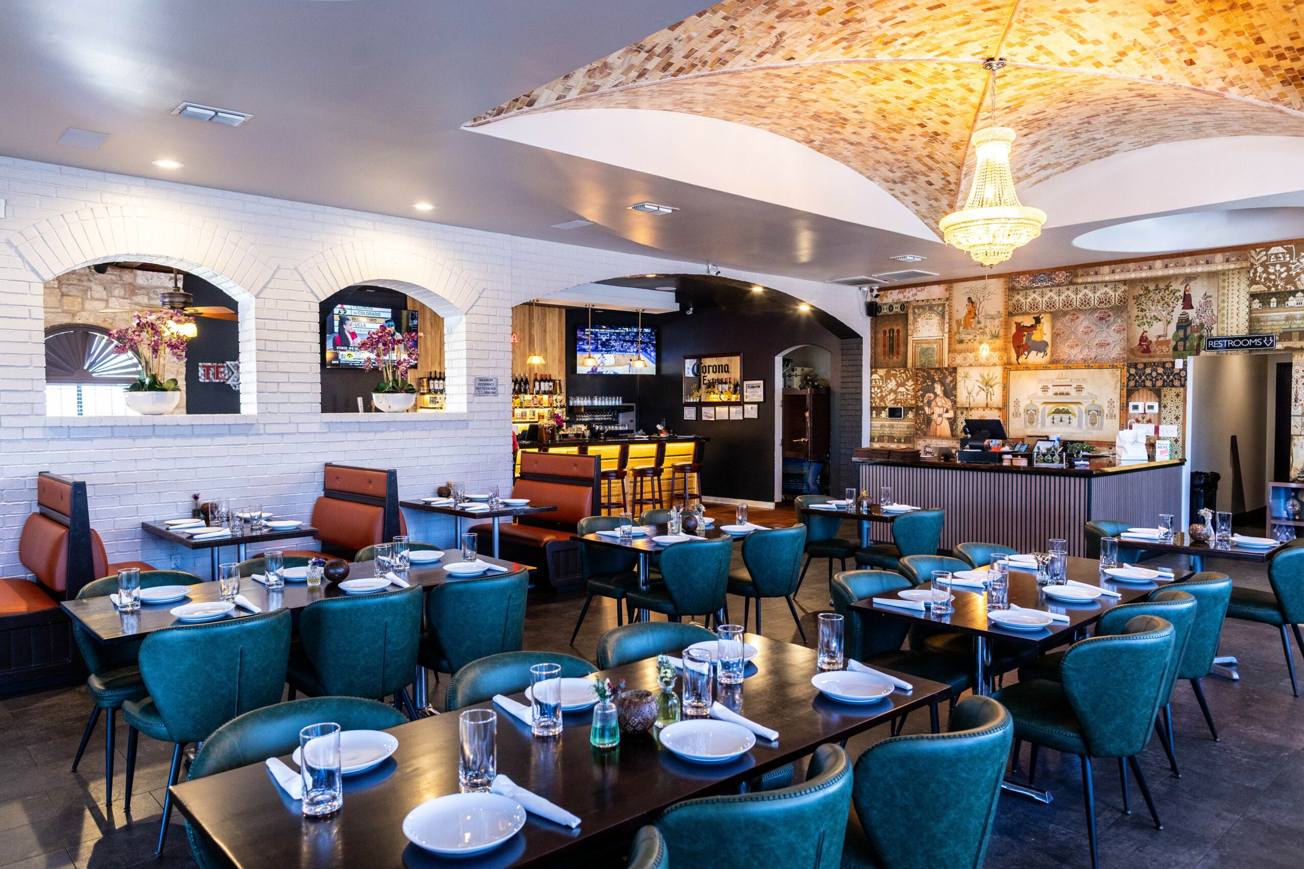 Restaurant interior with blue chairs, tables set with glasses and plates, bar area with shelves, and decorative ceiling light.