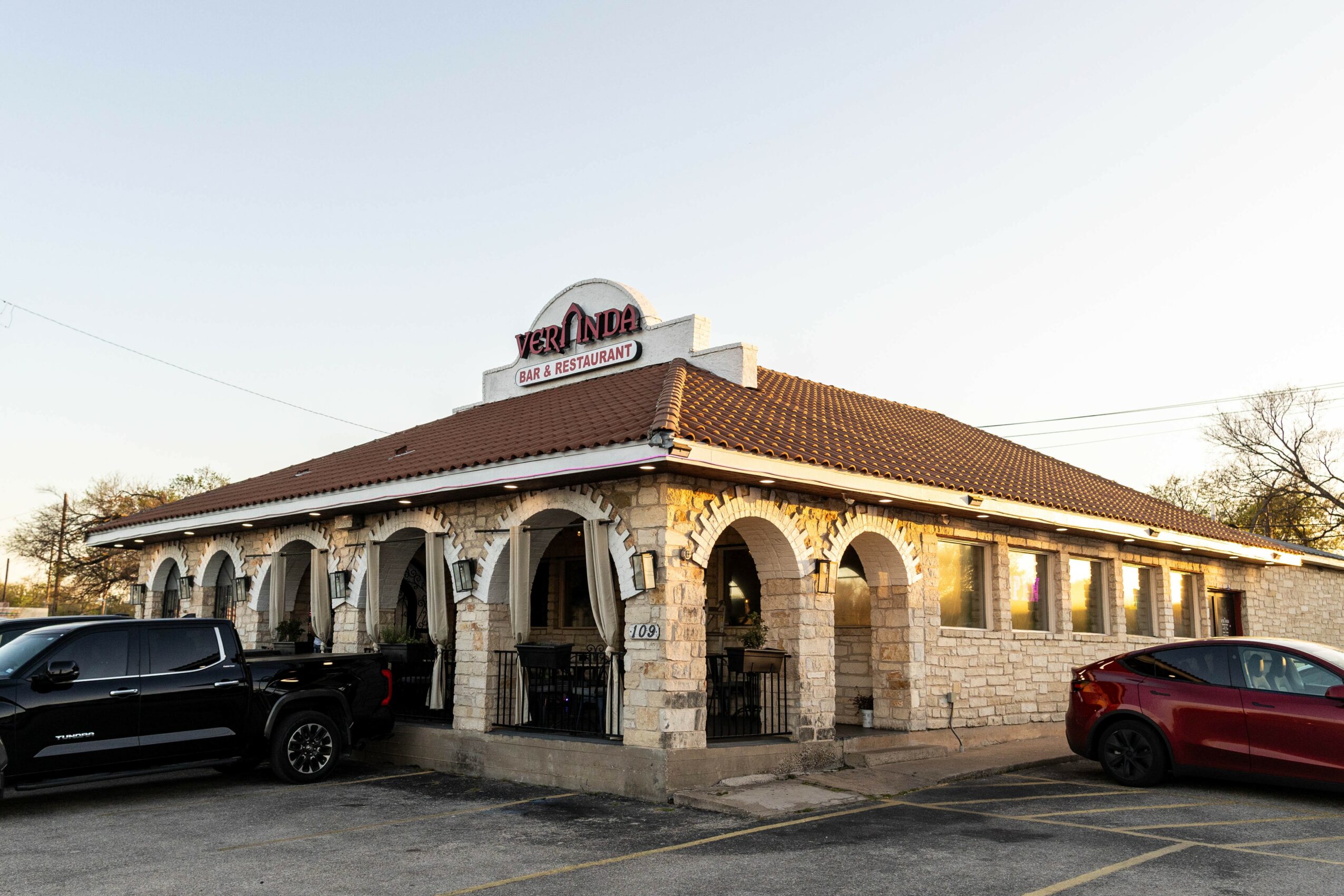 A single-story building with a red-tiled roof and stone archways, parked cars in front, and a sign on top, under a clear sky.