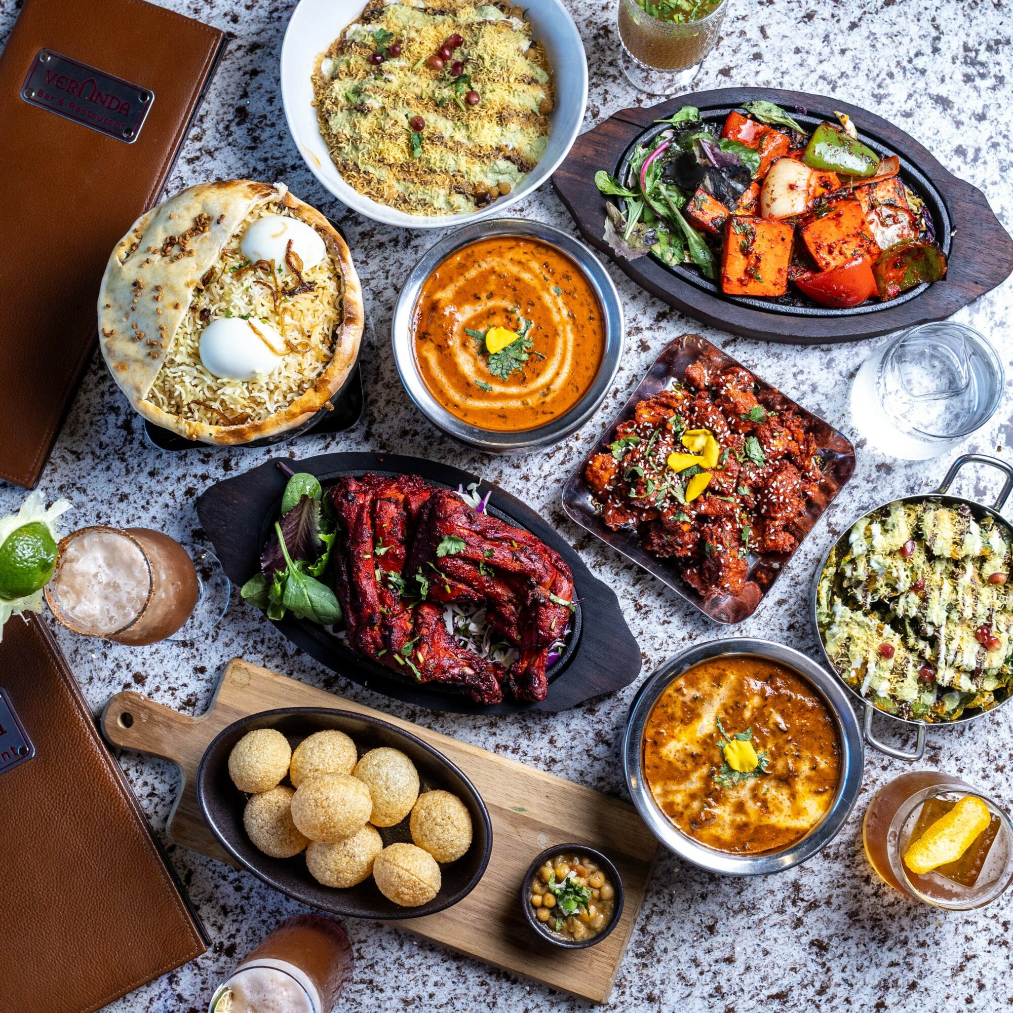 Assorted dishes including salad, curry, bread, and snacks on a speckled table with drinks and utensils.