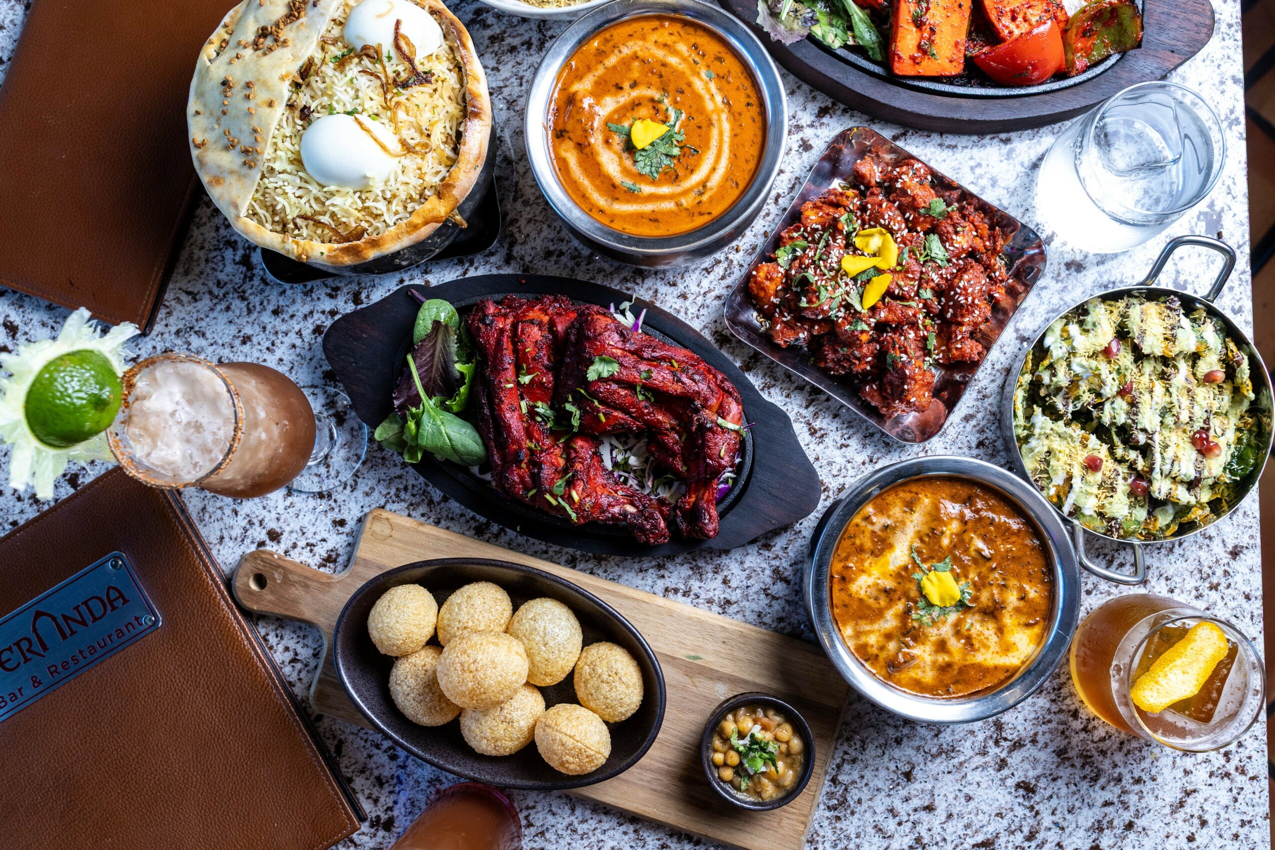 Assorted Indian dishes including naan, curry, grilled meat, rice, and drinks on a table.