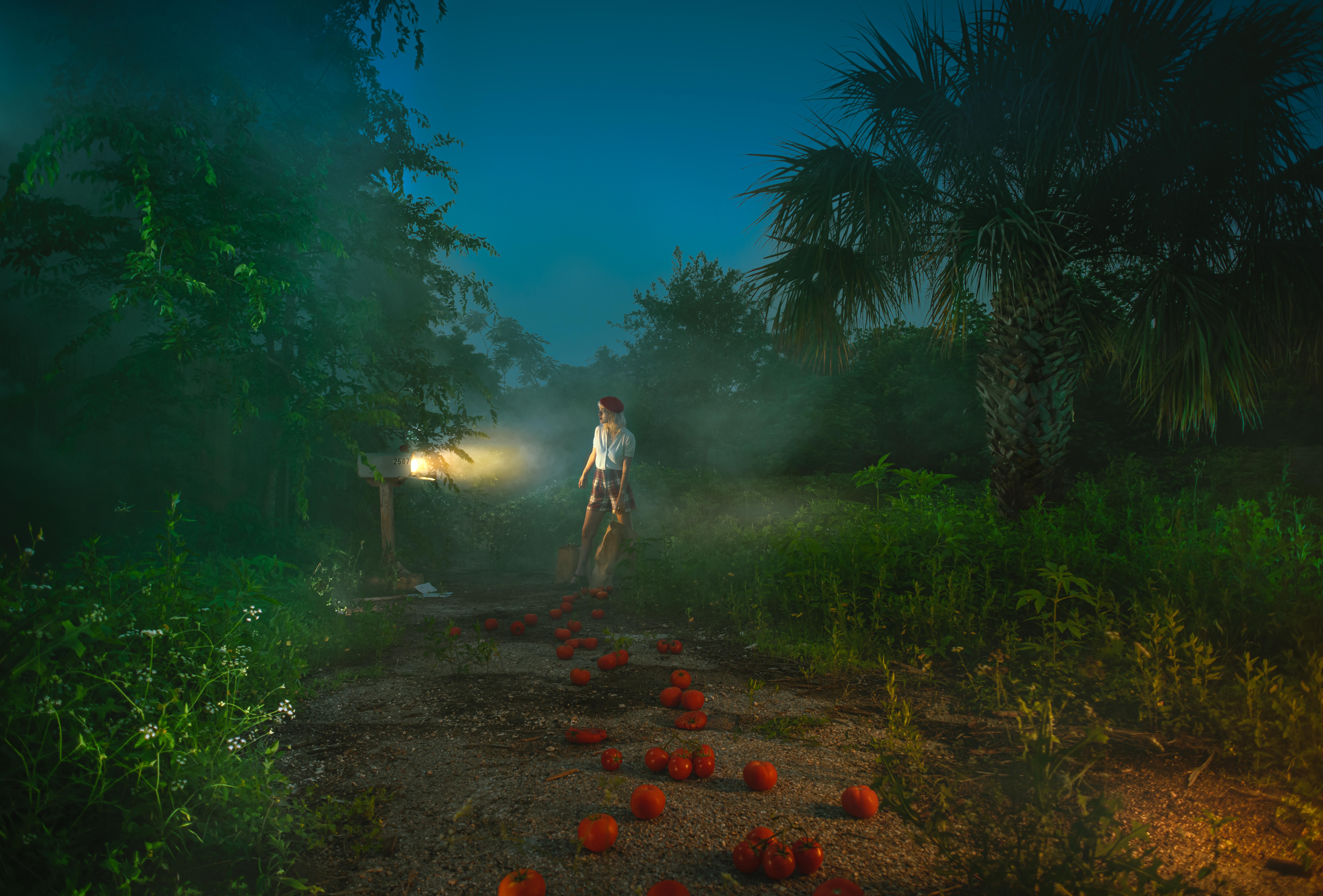 Person walking on a dirt path with scattered orange objects, surrounded by trees and foggy atmosphere at dusk.