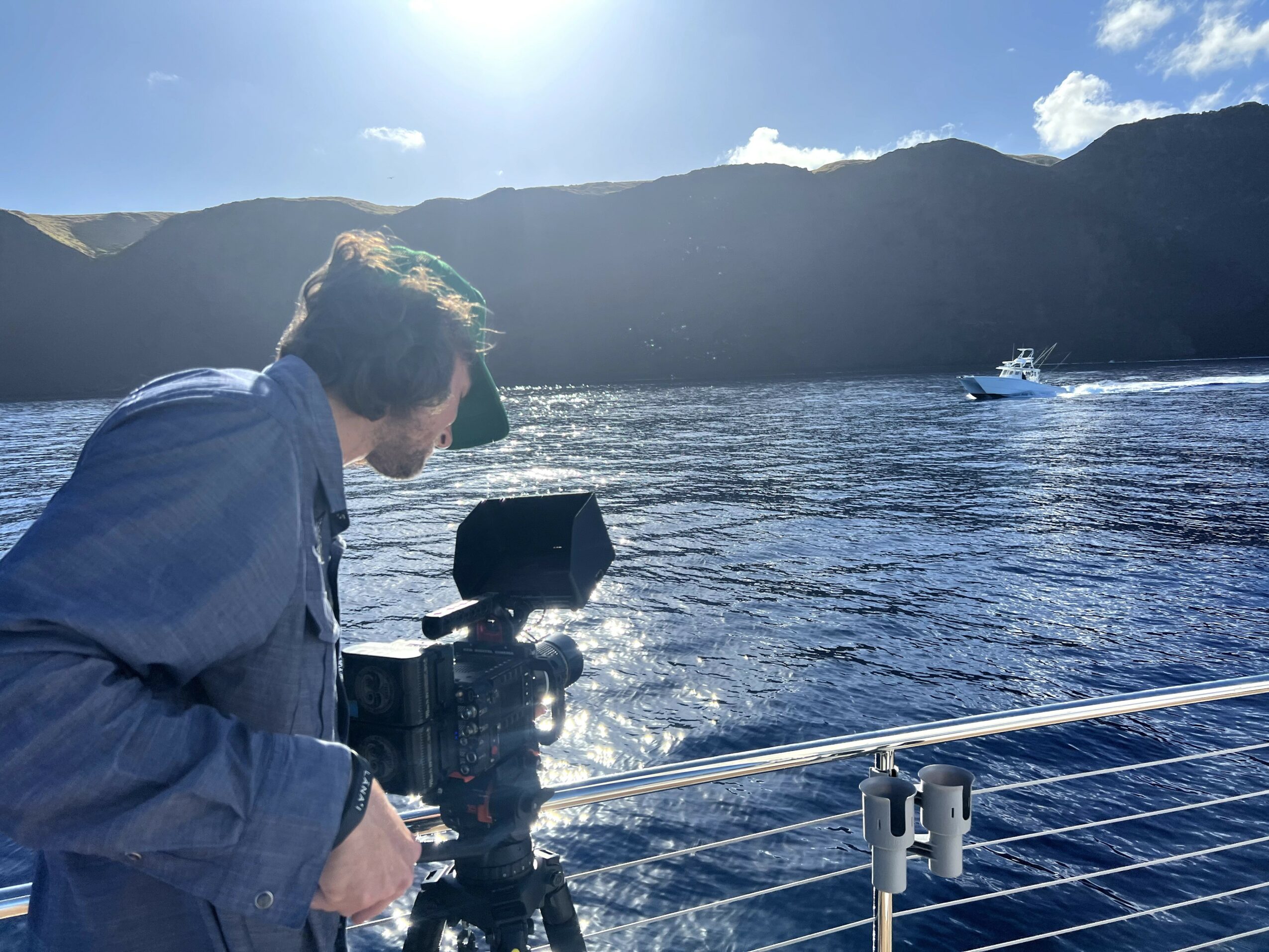 Person with camera on boat, water and mountains in background, sunny sky, and another boat in distance.