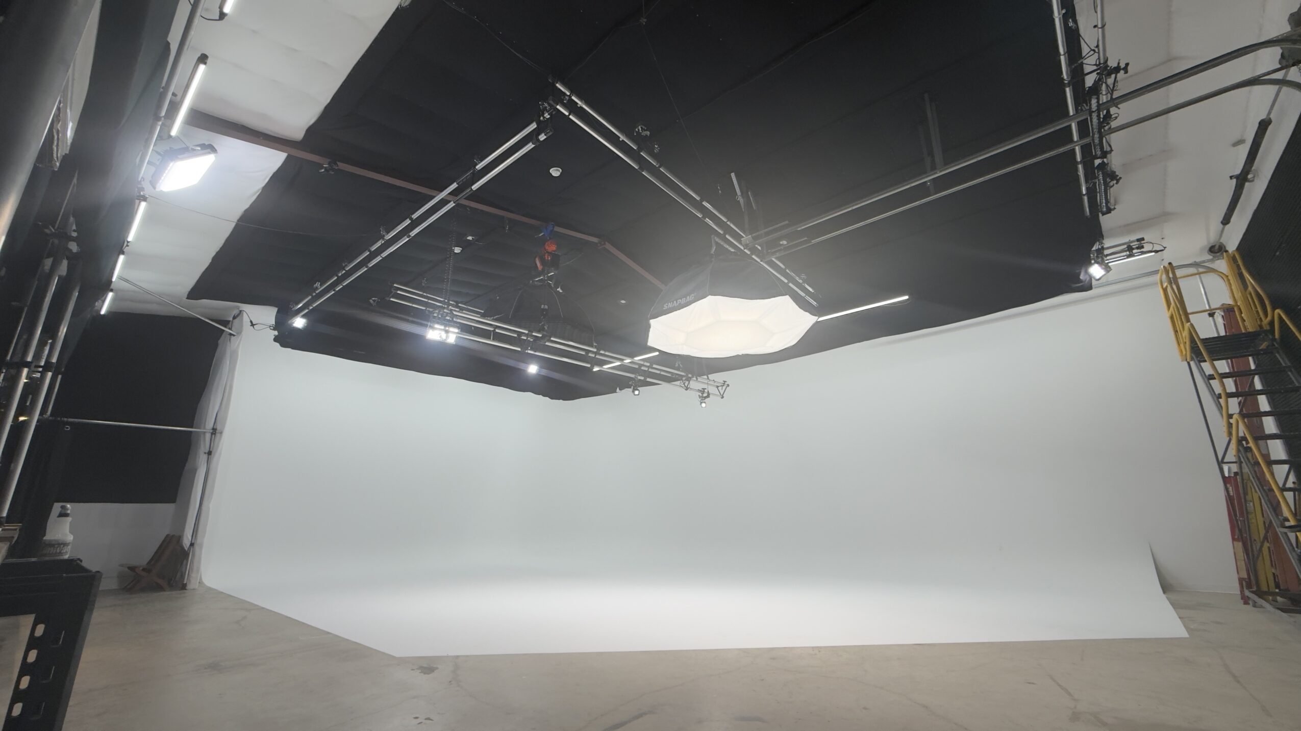 Studio with a large white backdrop and lighting equipment, ceiling with black panels, and a ladder on the right side.