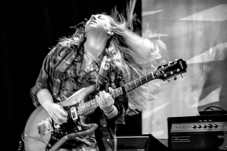 Woman playing electric guitar with hair flowing, stage background, black and white, energetic performance.