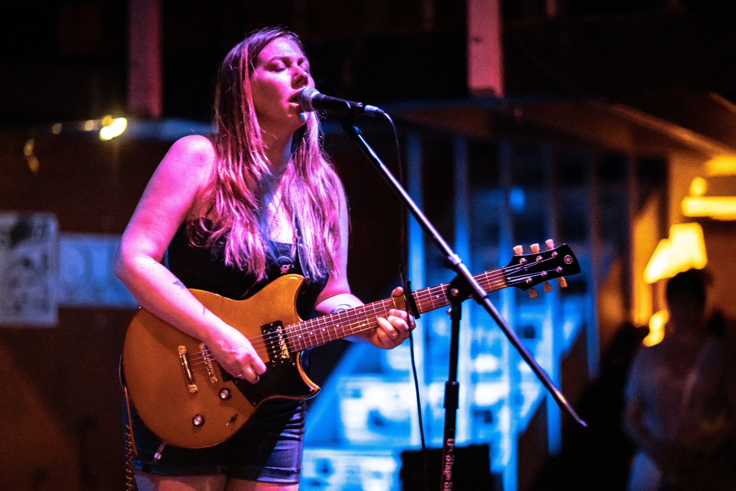 Female musician playing guitar and singing into microphone on stage with colorful lighting