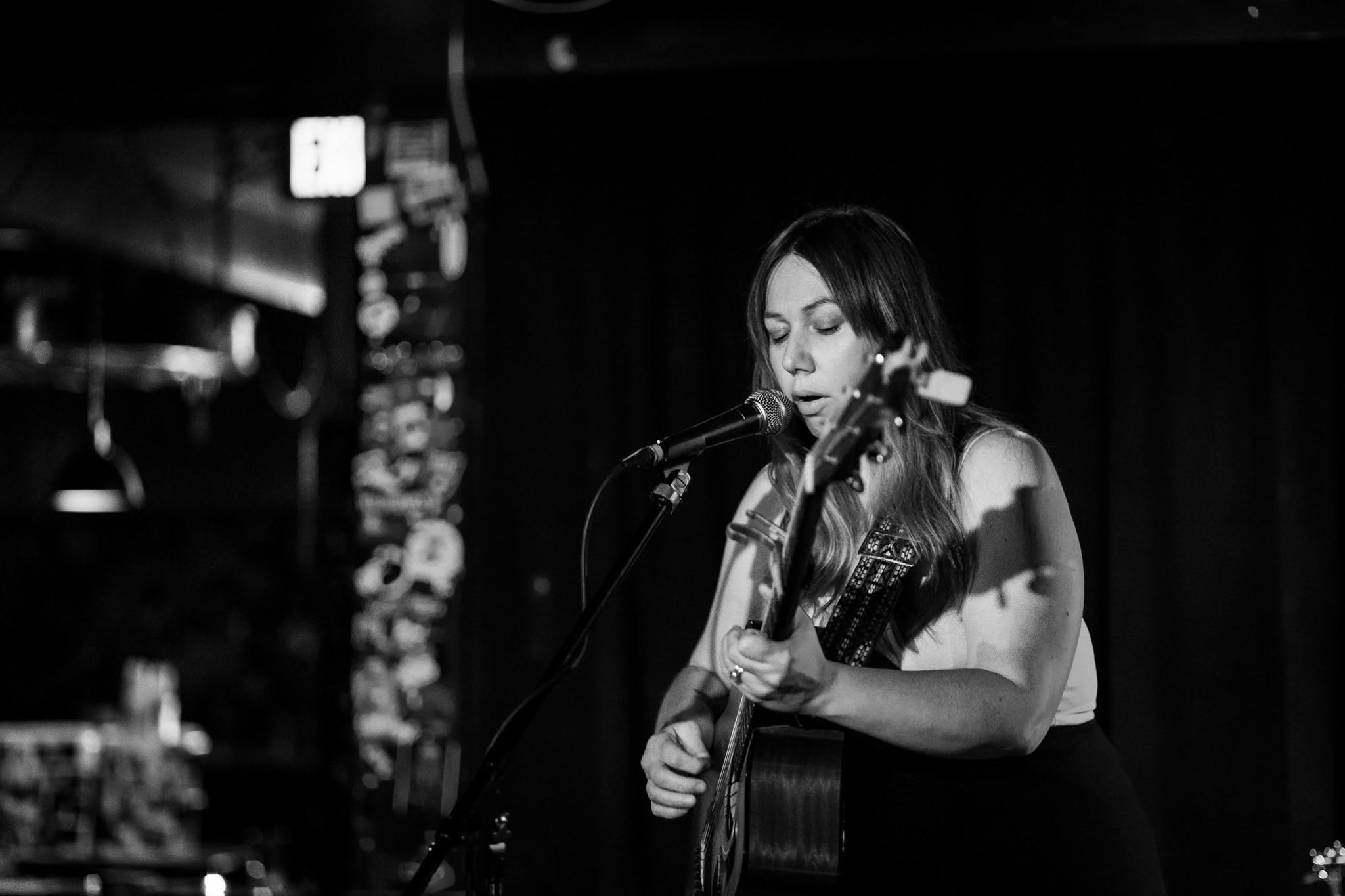 Woman playing guitar and singing into microphone on stage with dark background, black and white image.
