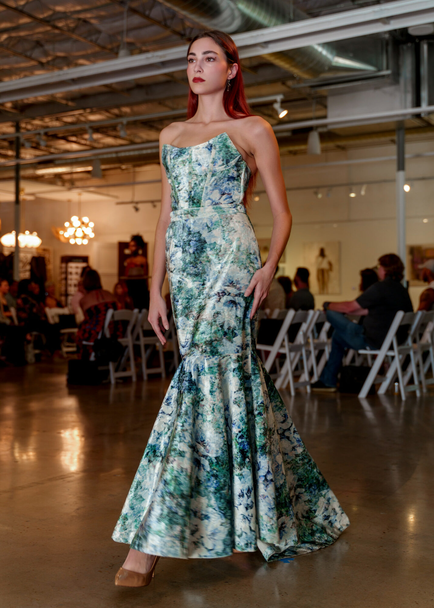 Woman in a strapless, floor-length floral gown standing in a decorated indoor event space with seated guests.