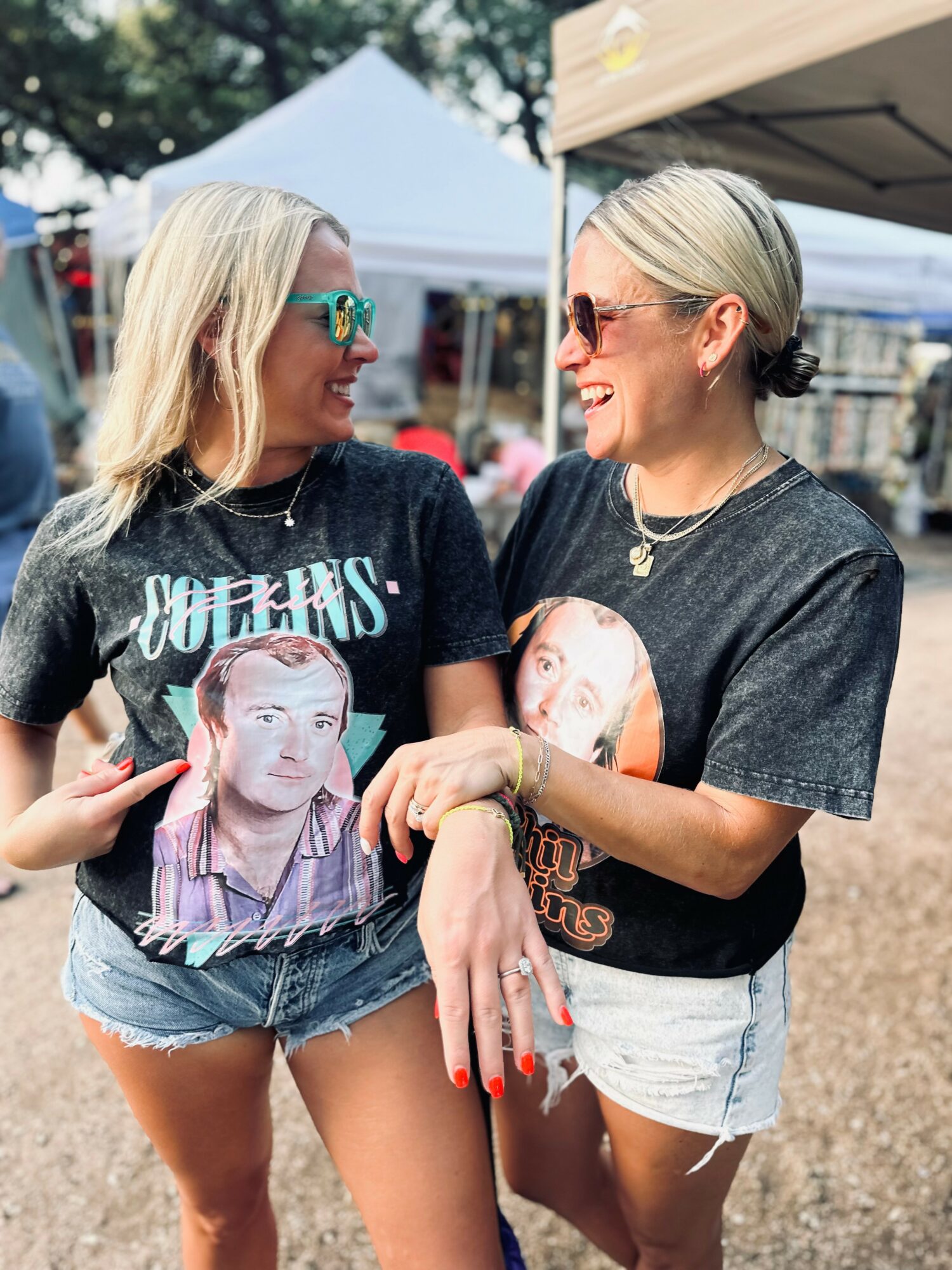 Two women with blonde hair and sunglasses smiling and talking outdoors at a festival or fair, wearing graphic t-shirts and shorts.