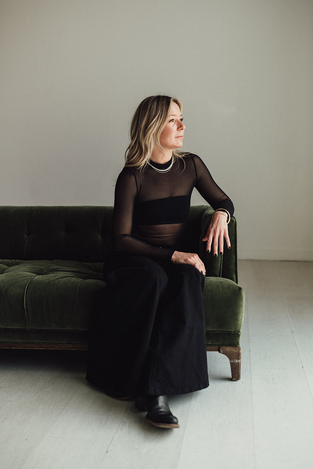 Woman with shoulder-length hair sitting on a dark green sofa, wearing a black sheer top and long black skirt, looking to the side.