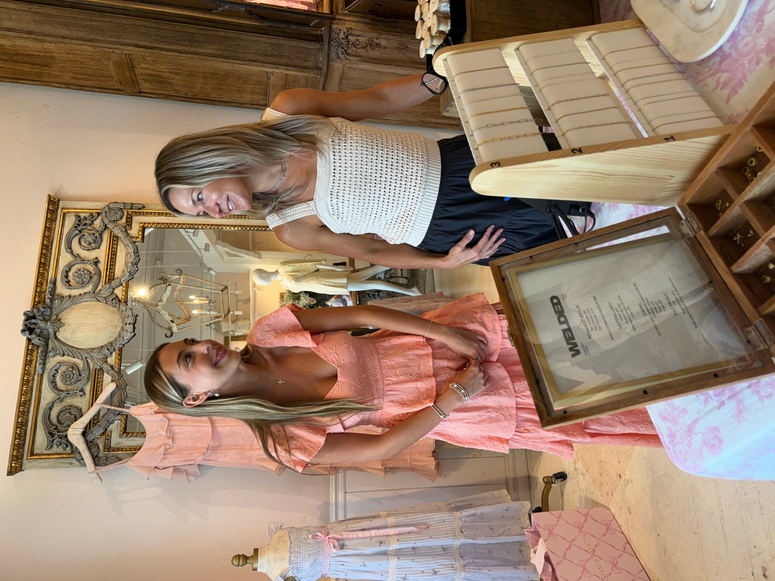 Two women standing in a room with ornate decor, one in a white top and the other in a pink dress, near a framed document.
