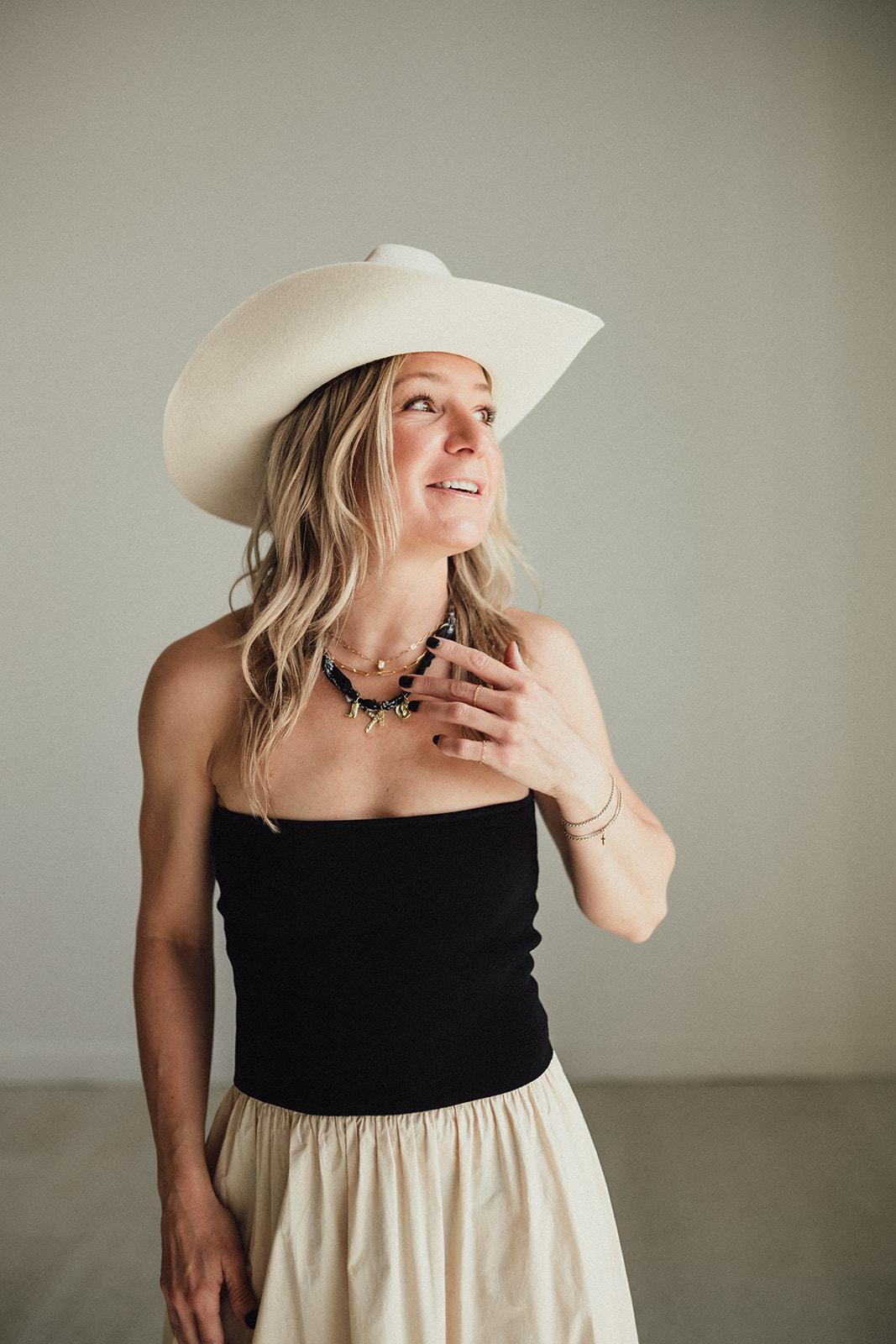 Woman wearing a large white hat, black strapless top, and beige skirt, looking upward with a smile, holding her necklace.