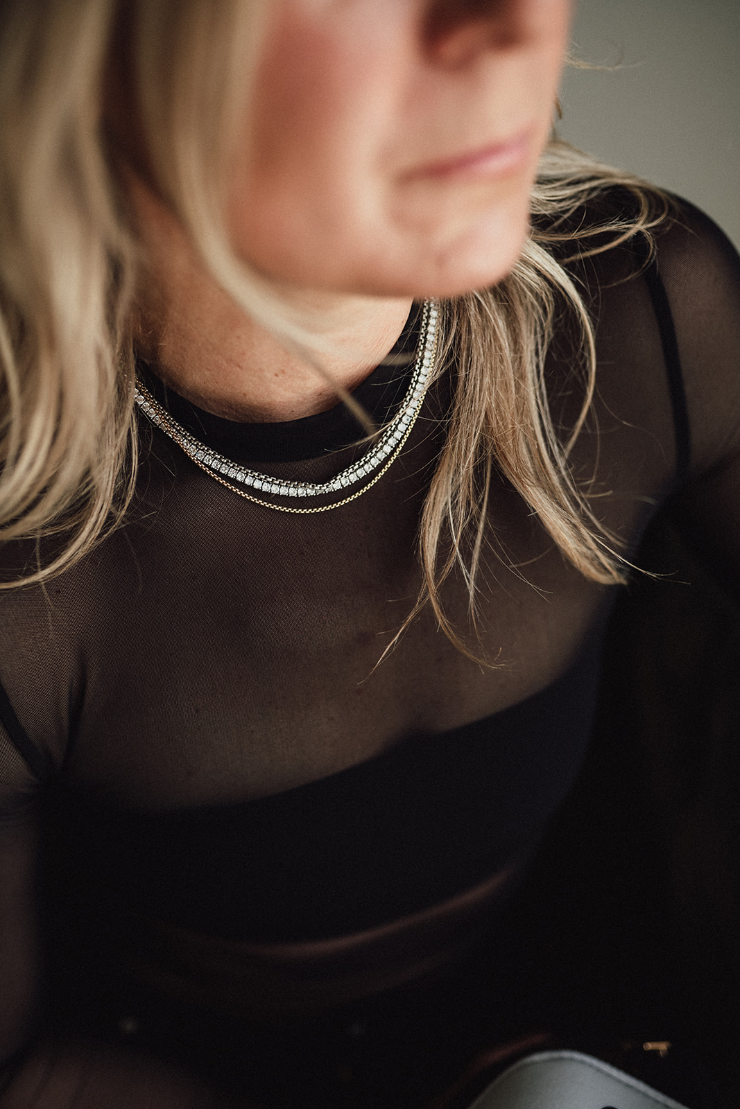 Close-up of a woman with blonde hair wearing layered necklaces and a black sheer top.