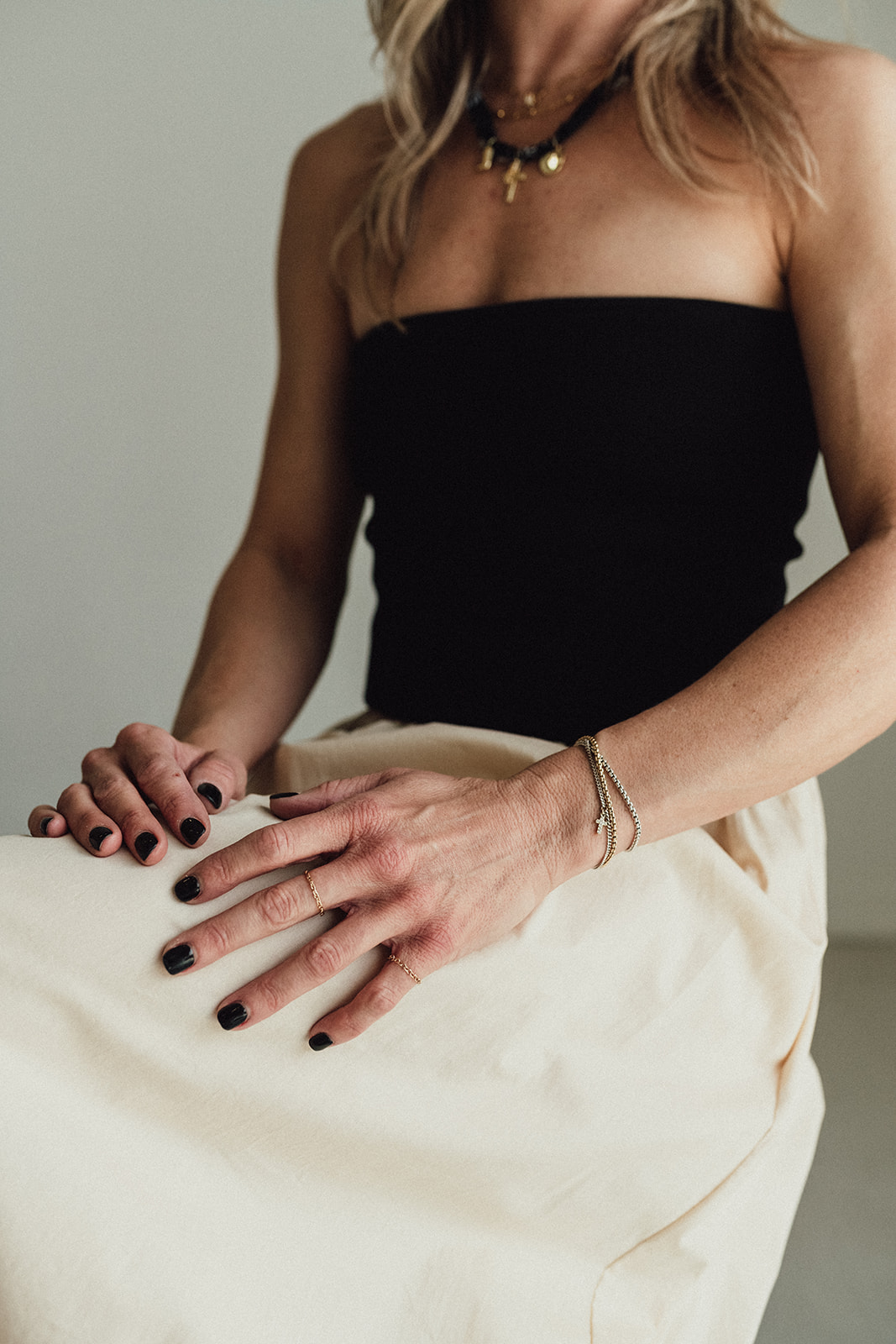 Close-up of a woman wearing a black strapless top, with hands resting on her lap, showing dark nail polish and jewelry.