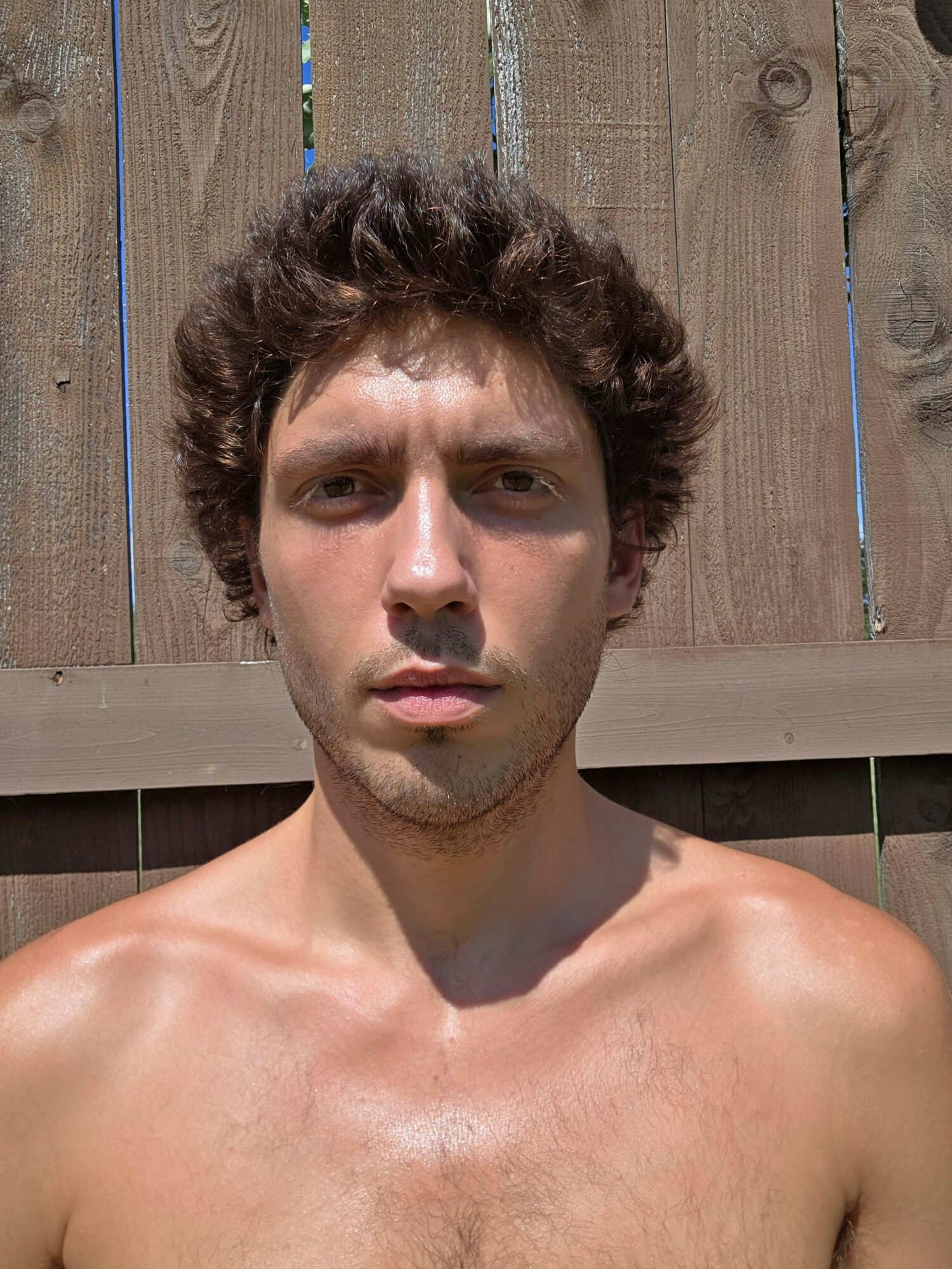Young man with curly hair and bare shoulders outdoors, standing in front of a wooden fence.