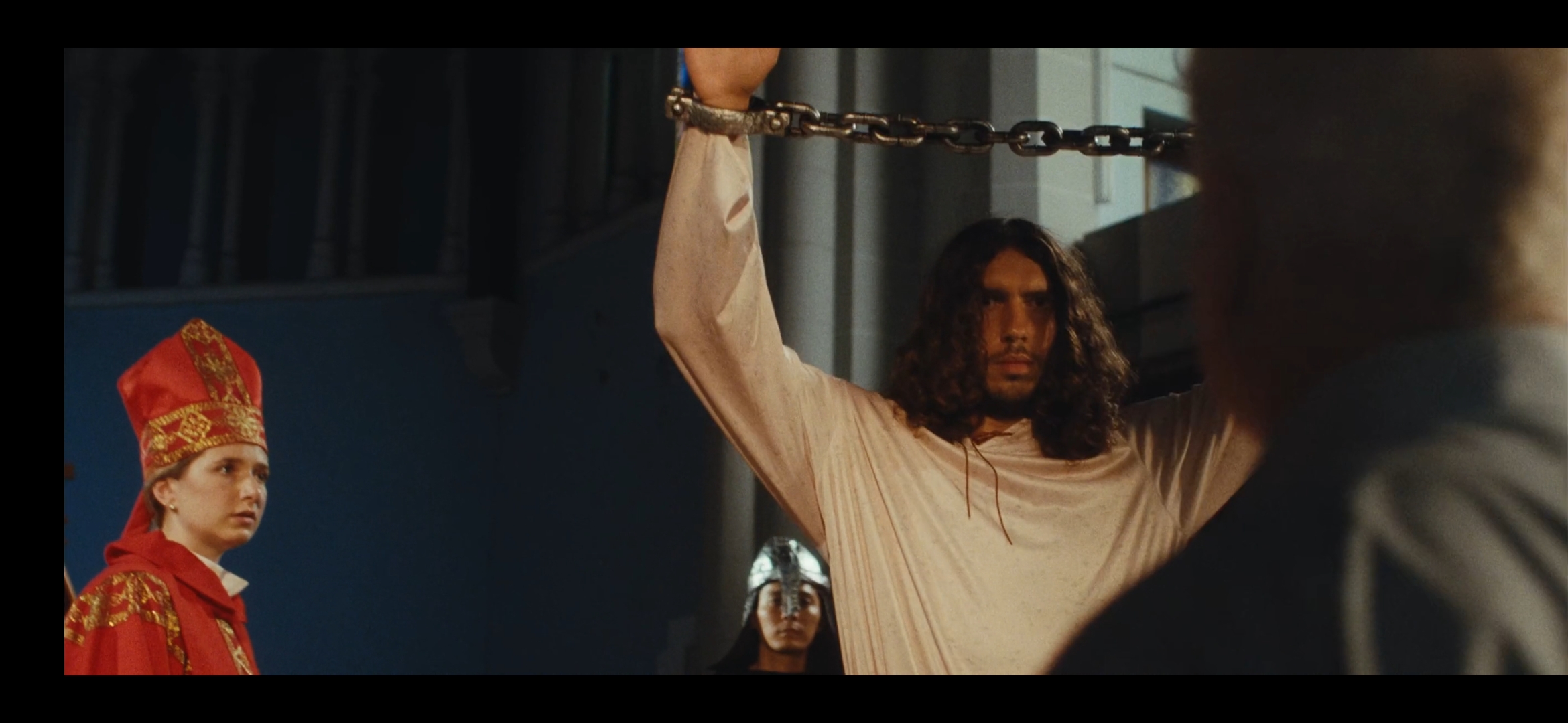 Person with long hair and light-colored clothing holding a chain overhead, facing another person with short hair, in a room with a blue background and a woman in traditional attire. Word count: 20