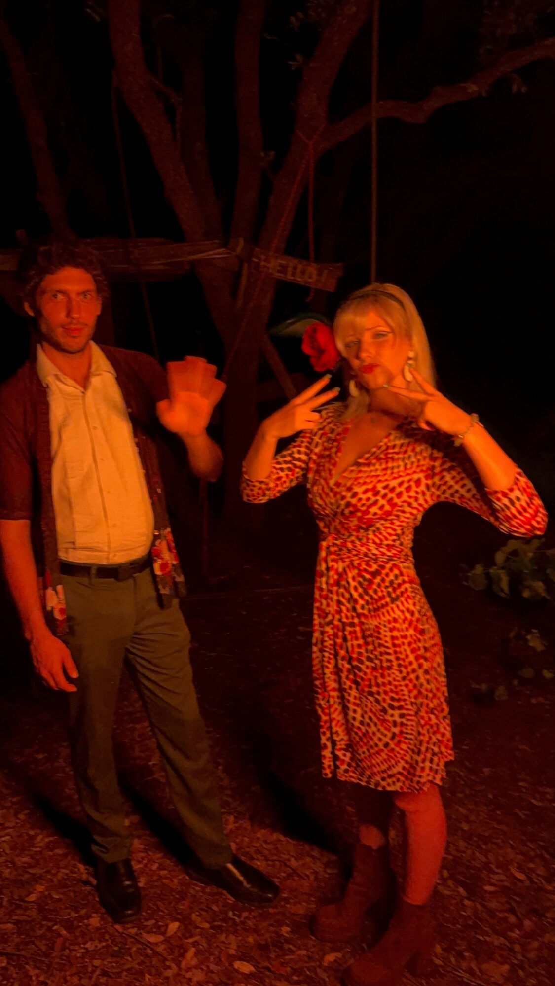 Man and woman standing outdoors at night, woman drinking from a glass, tree in background, dim lighting.