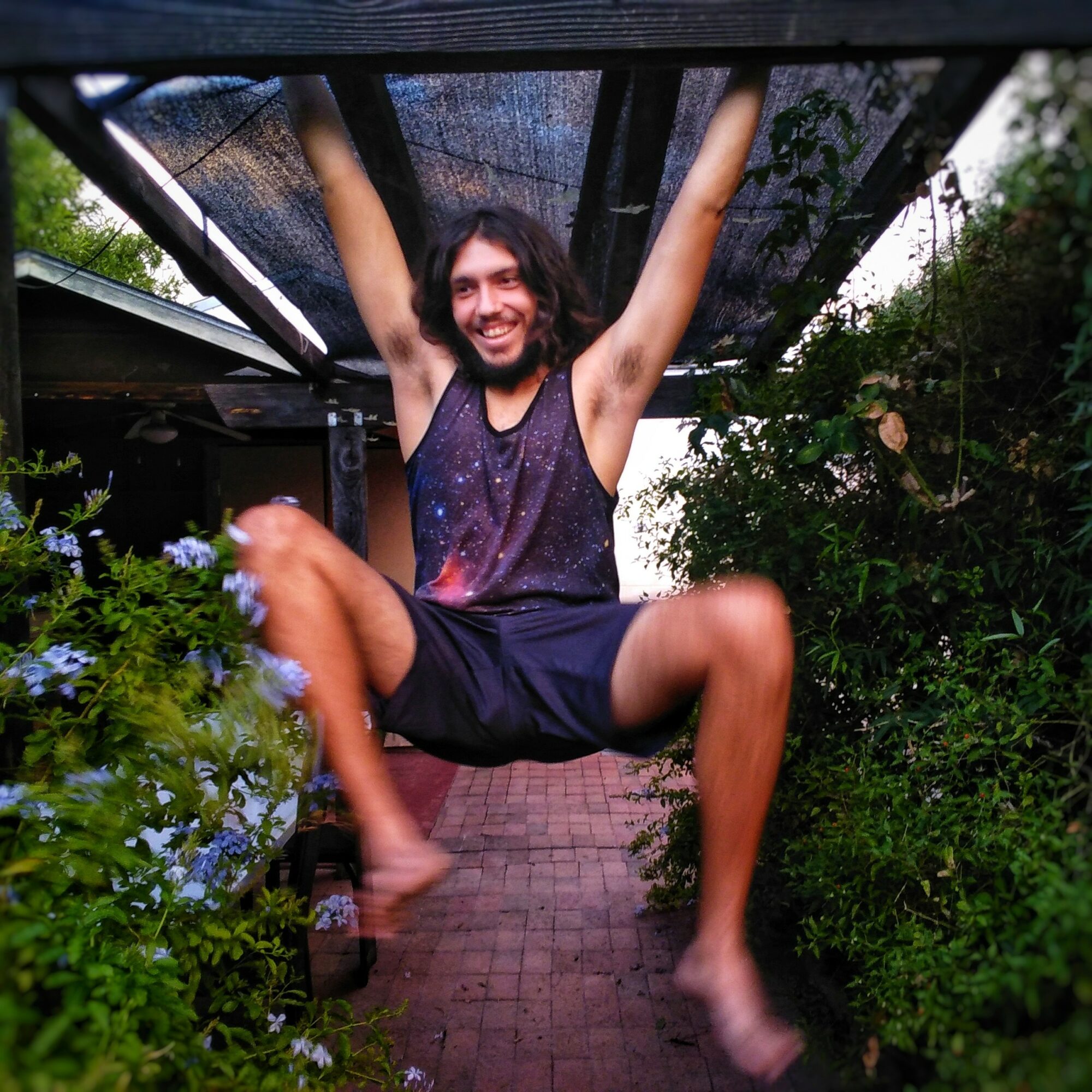 Person hanging upside down from a structure, smiling, surrounded by greenery and flowers, with arms raised and legs bent.