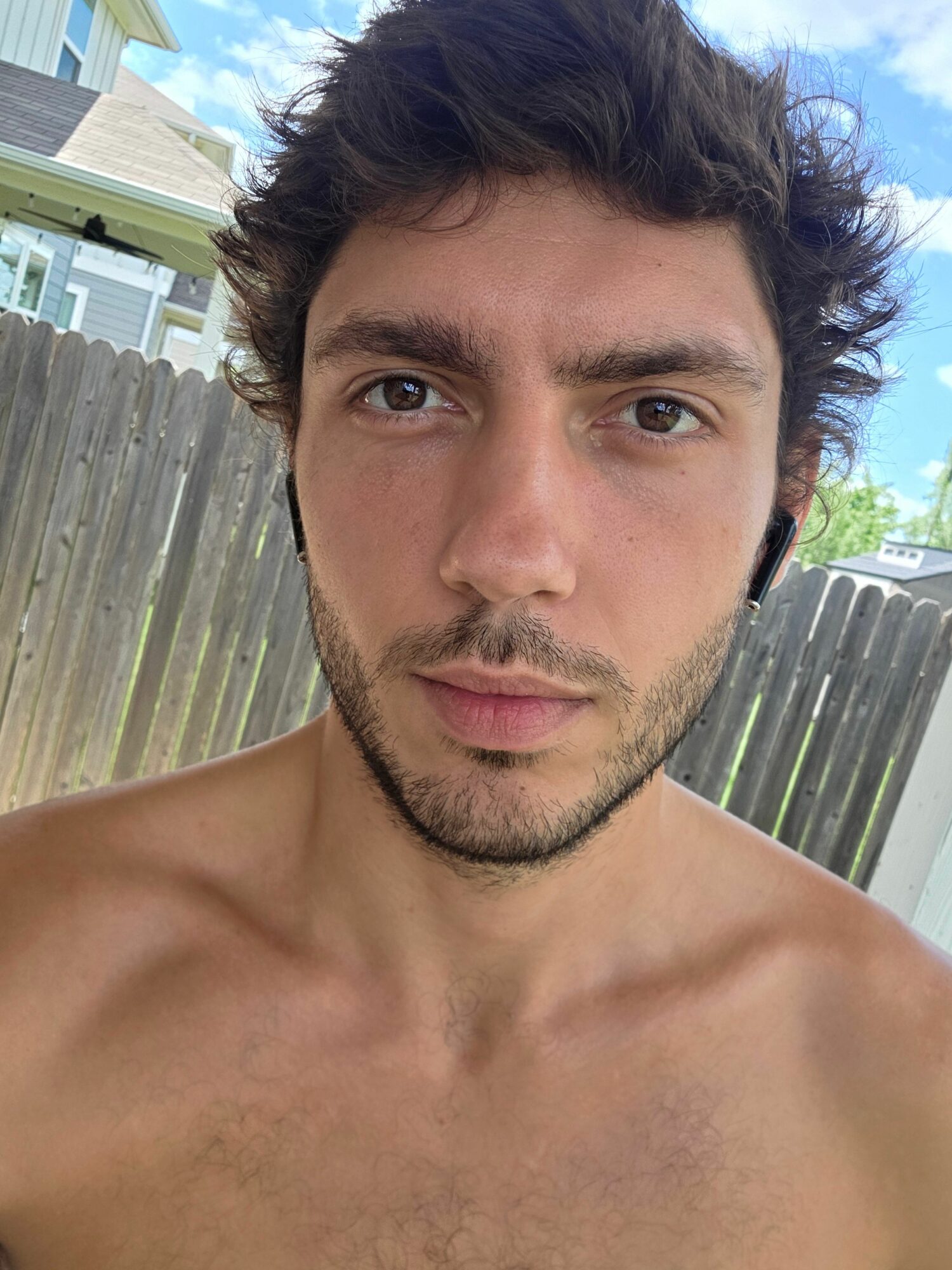 A shirtless man with dark, wavy hair and light eyes looks at the camera outdoors, with a wooden fence and blue sky in background.