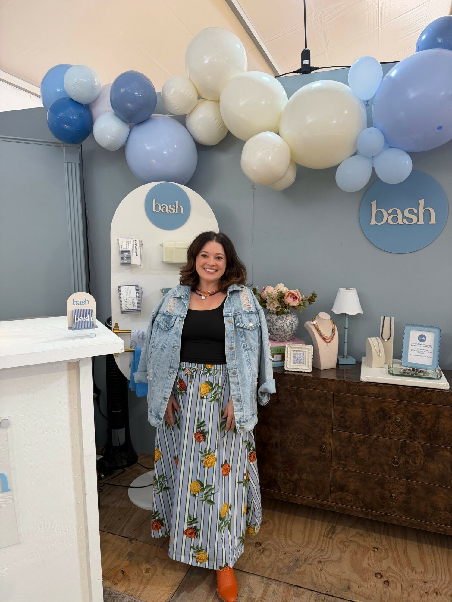 Woman standing in front of a decorated wall with balloons and a table, smiling, wearing a black top and a floral skirt.